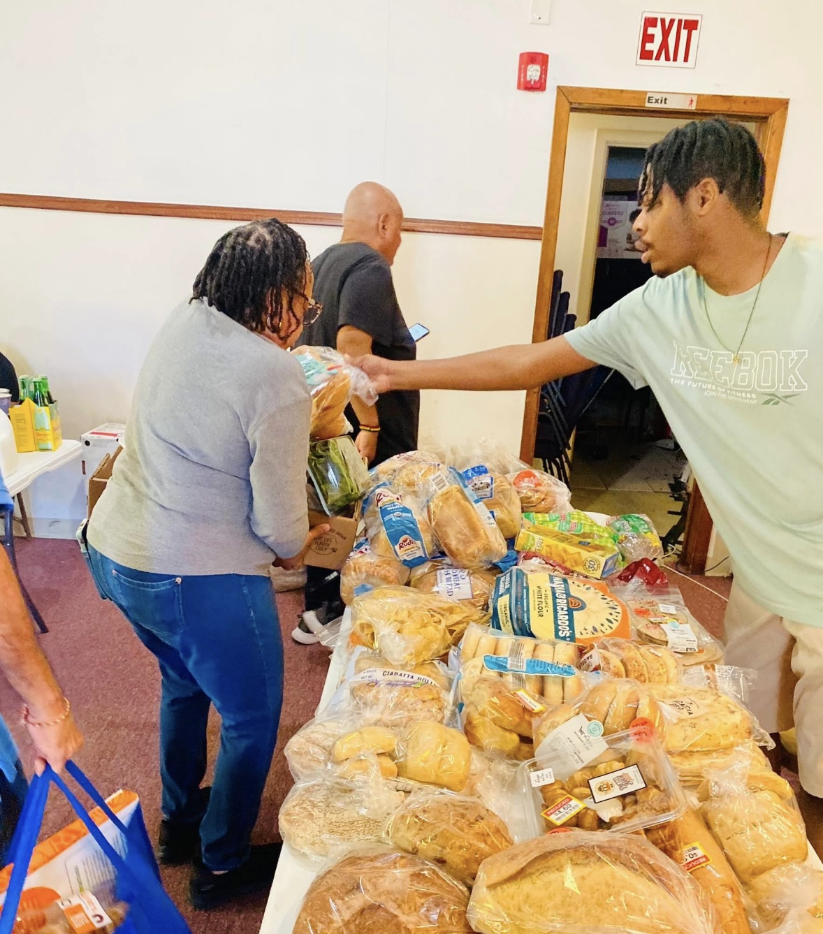 Young man hands bread to woman at table with bread and snacks, in a room with an exit sign above door.