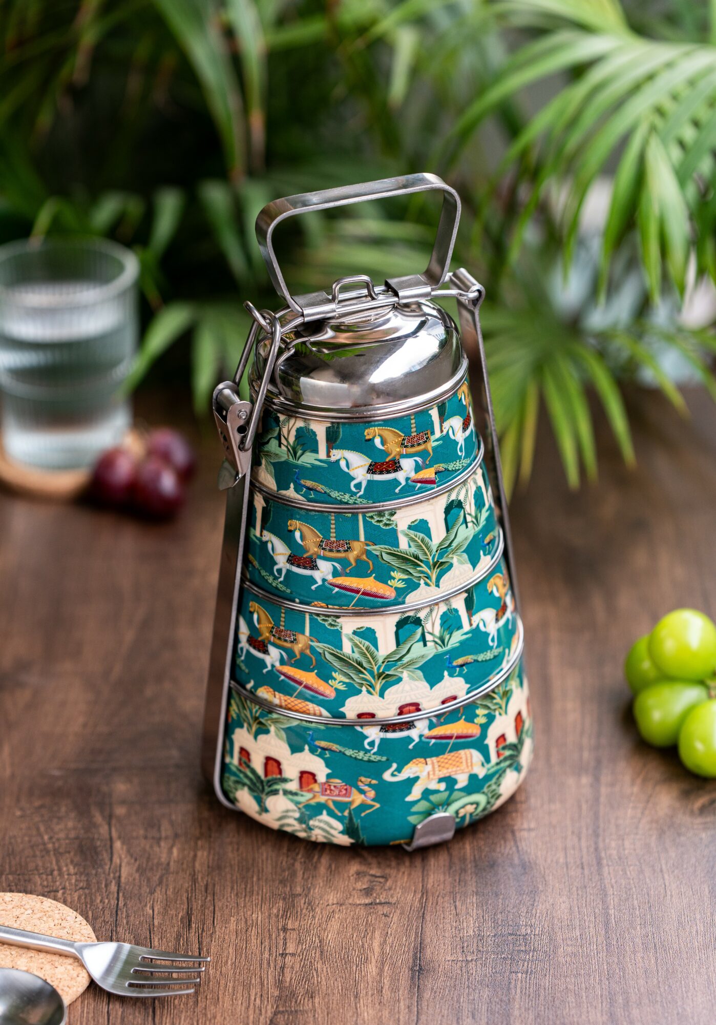 A metal kettle with a colorful patterned fabric cover featuring animals and plants, placed on a wooden table with green plants in background.