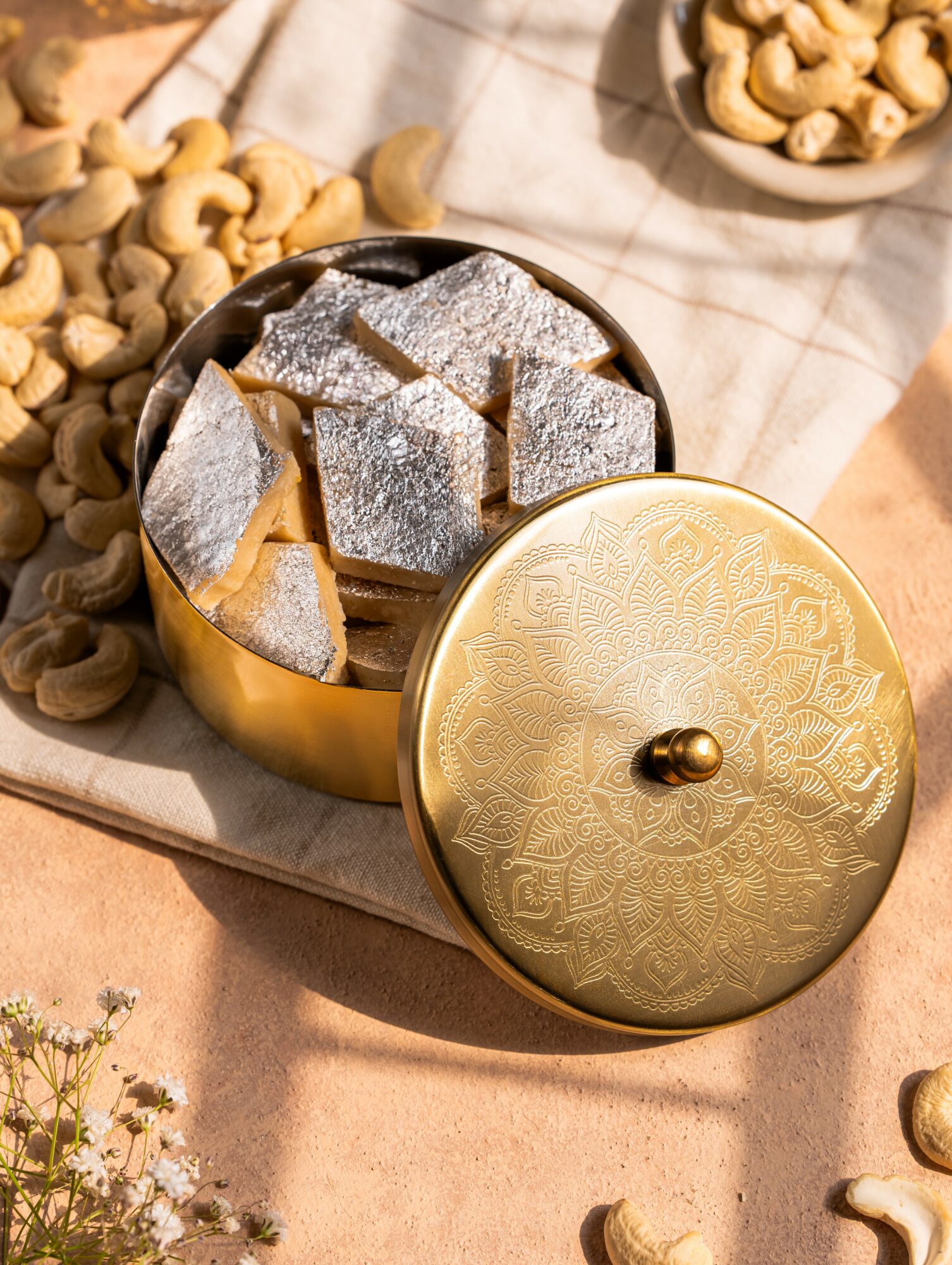 Golden container with lid open, filled with sugar cubes, on a table with nuts and a cloth, sunlight casting shadows.