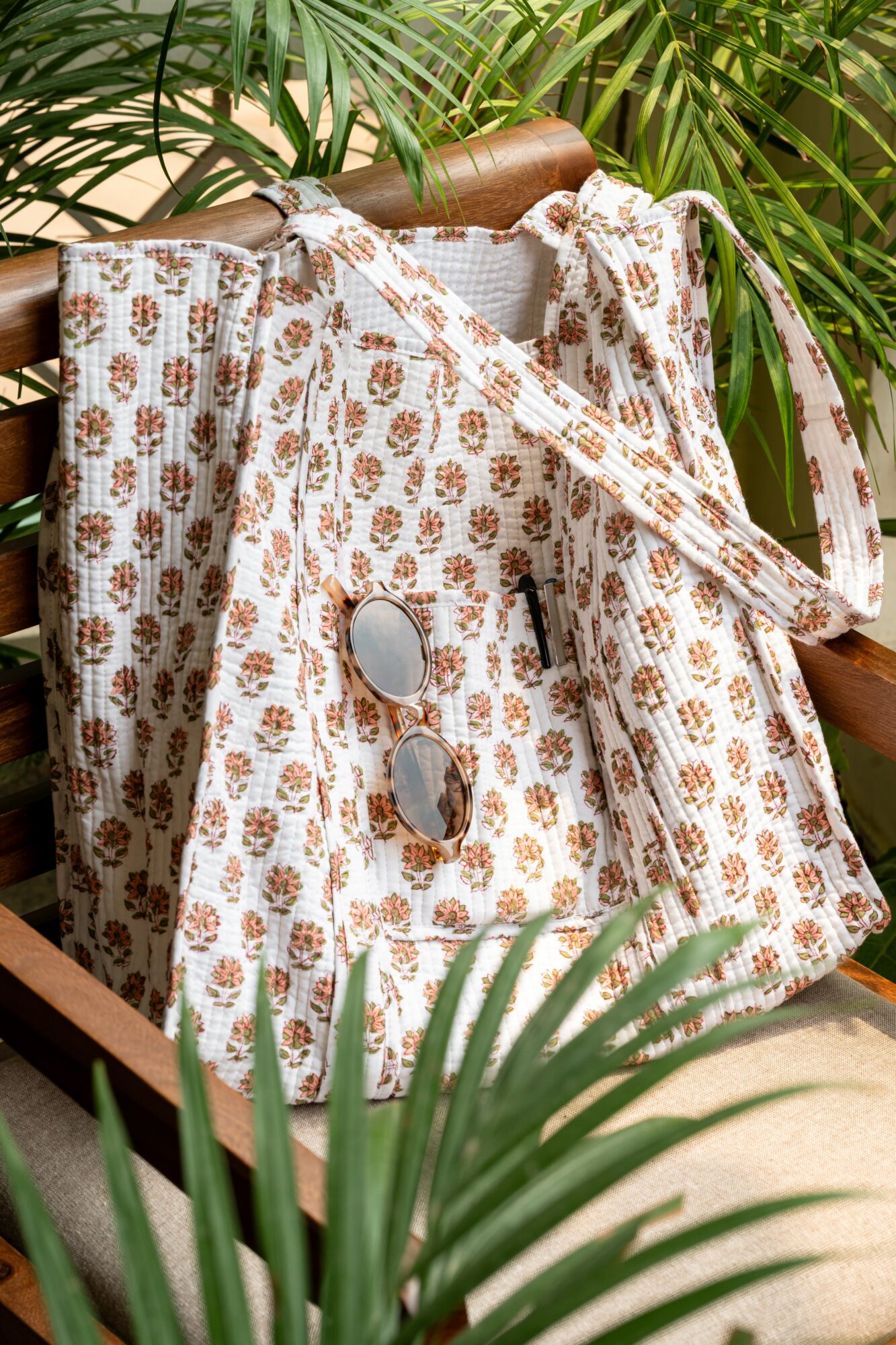 A patterned fabric bag with sunglasses and a pen, placed on a wooden chair surrounded by green plants.