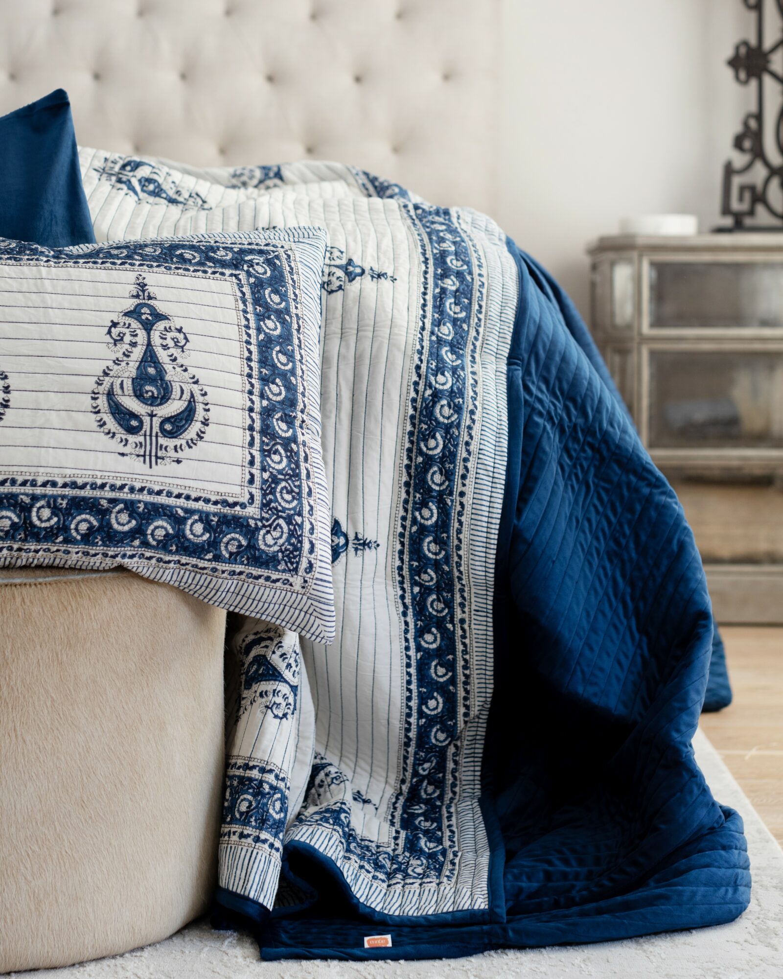 Close-up of a bed with a white tufted headboard, blue and white patterned quilt, and a blue blanket, in a bedroom.