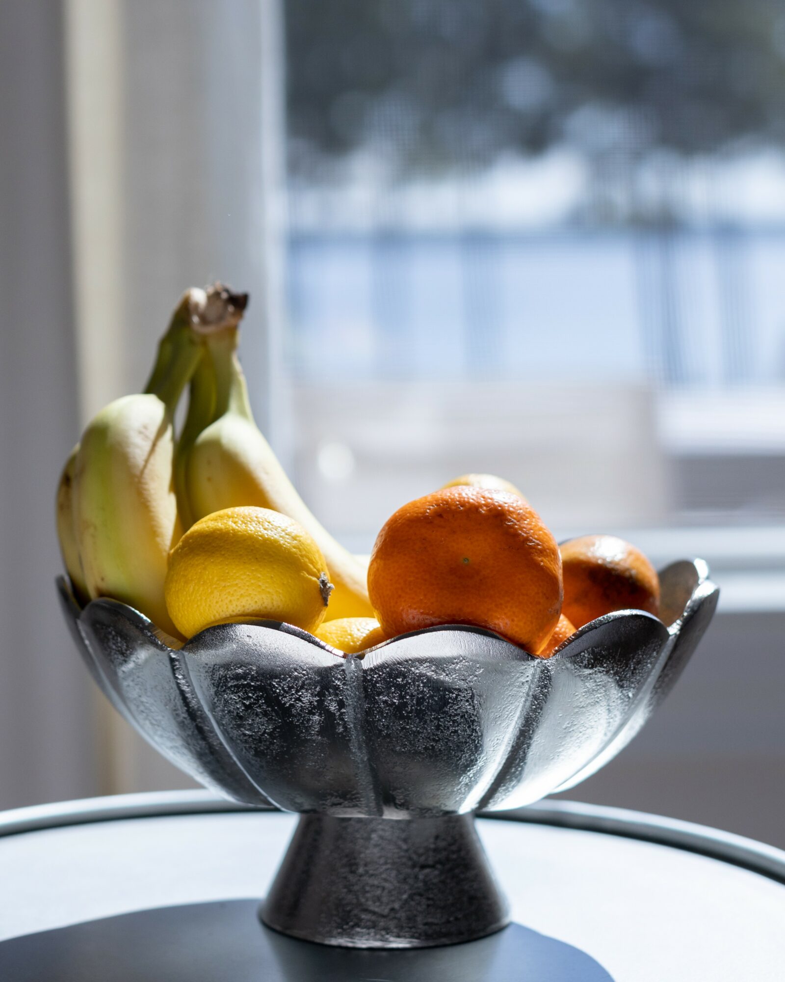 A glass bowl filled with bananas, an orange, and a lemon on a table near a window with blurred outdoor scenery.