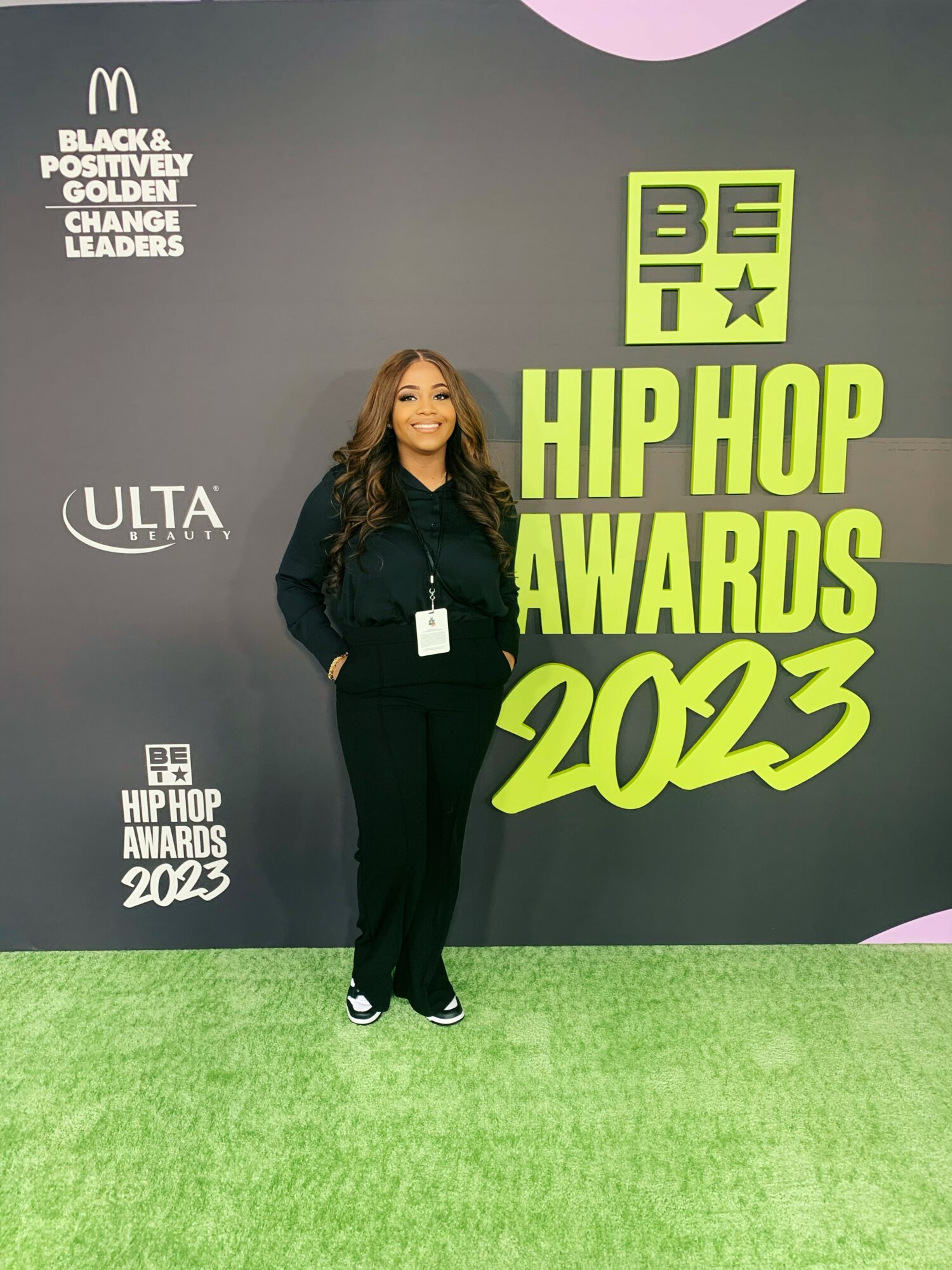 Woman standing on green carpet at hip hop awards event, backdrop with logos and text, smiling, wearing black outfit.