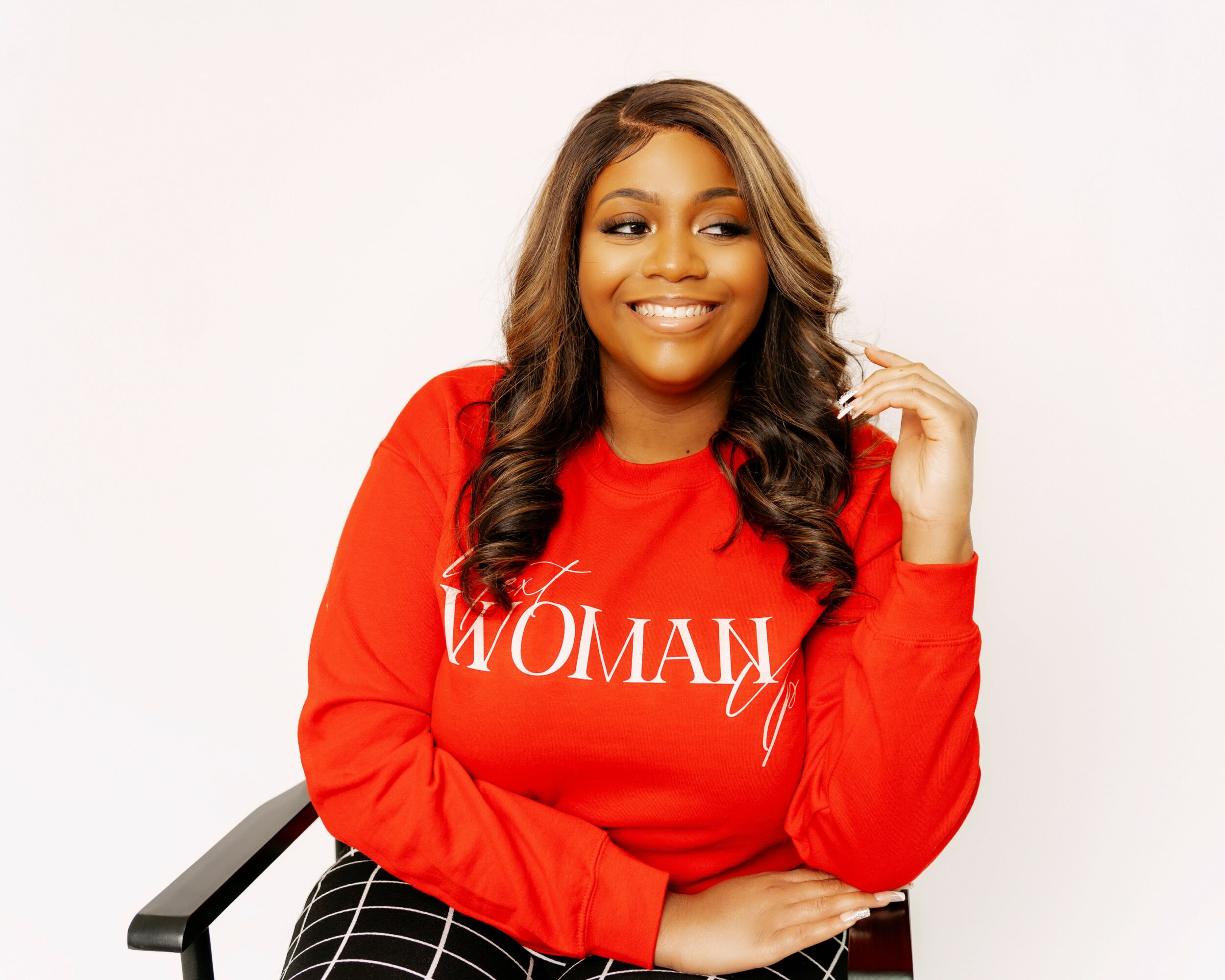 Smiling woman with long wavy hair wearing a red sweatshirt with white text, sitting on a chair against a white background.