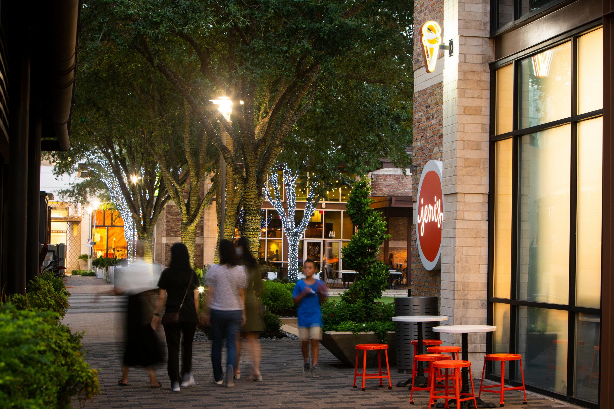 People walking on a sidewalk with trees and buildings, sunlight shining through trees, outdoor seating with red stools, and a large window.