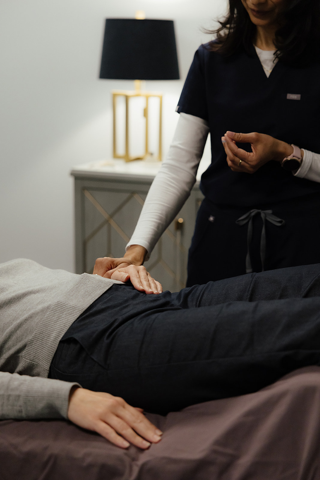 Healthcare professional examining a patient's abdomen in a clinical setting, with a bedside table and lamp in the background.