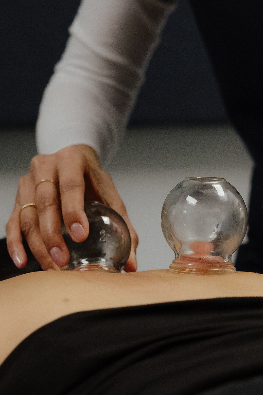 Person performing a cupping therapy with glass cups on their back, hand holding a cup, background is dark.