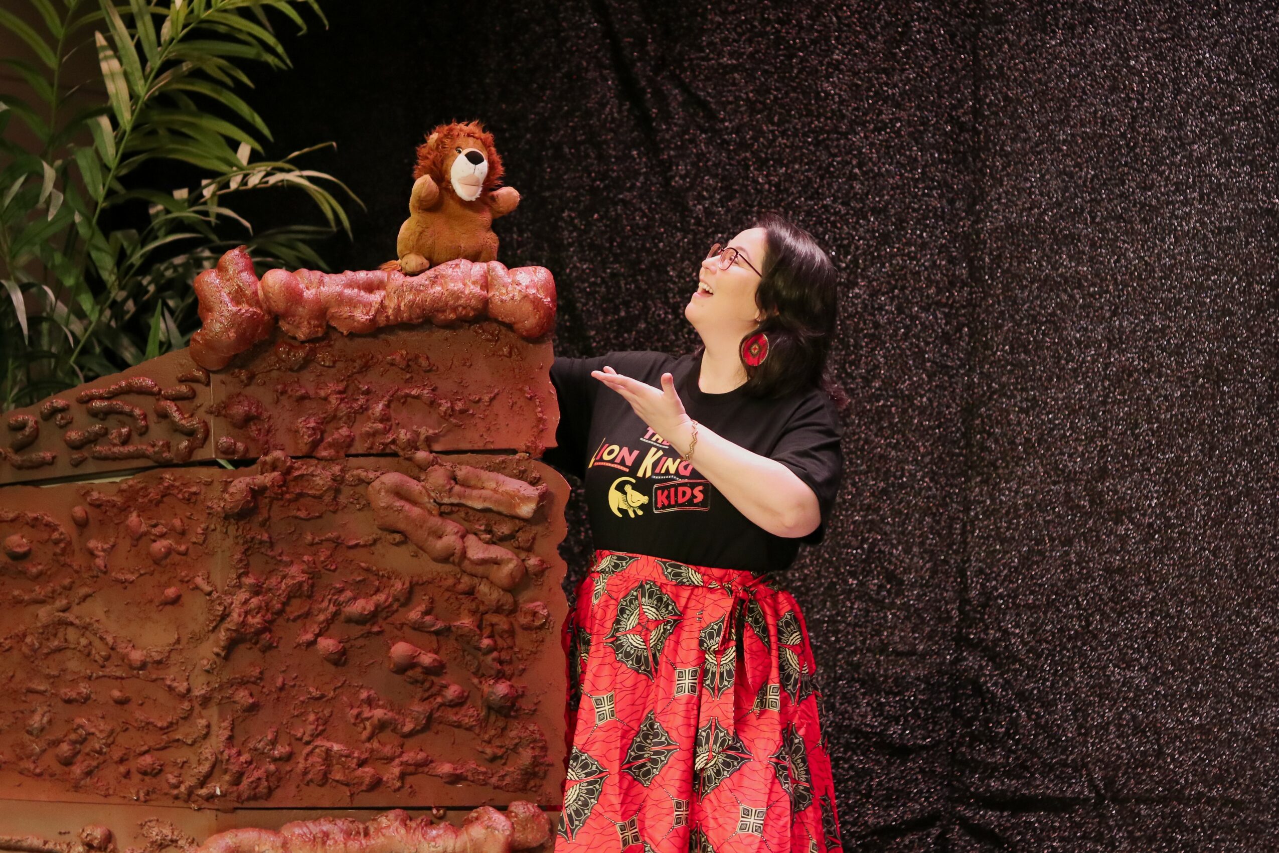 Person with black hair and glasses pointing at a lion puppet on a rock, with plants nearby.