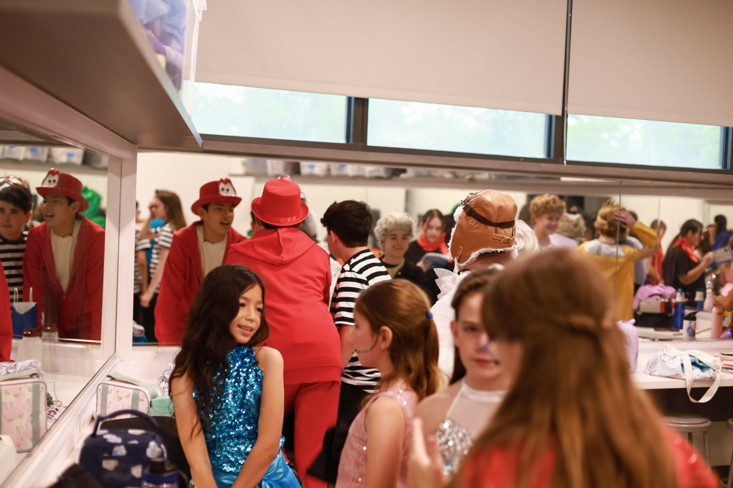 People gathered in a brightly lit room, some wearing colorful costumes and hats, with a mirror reflecting the scene.
