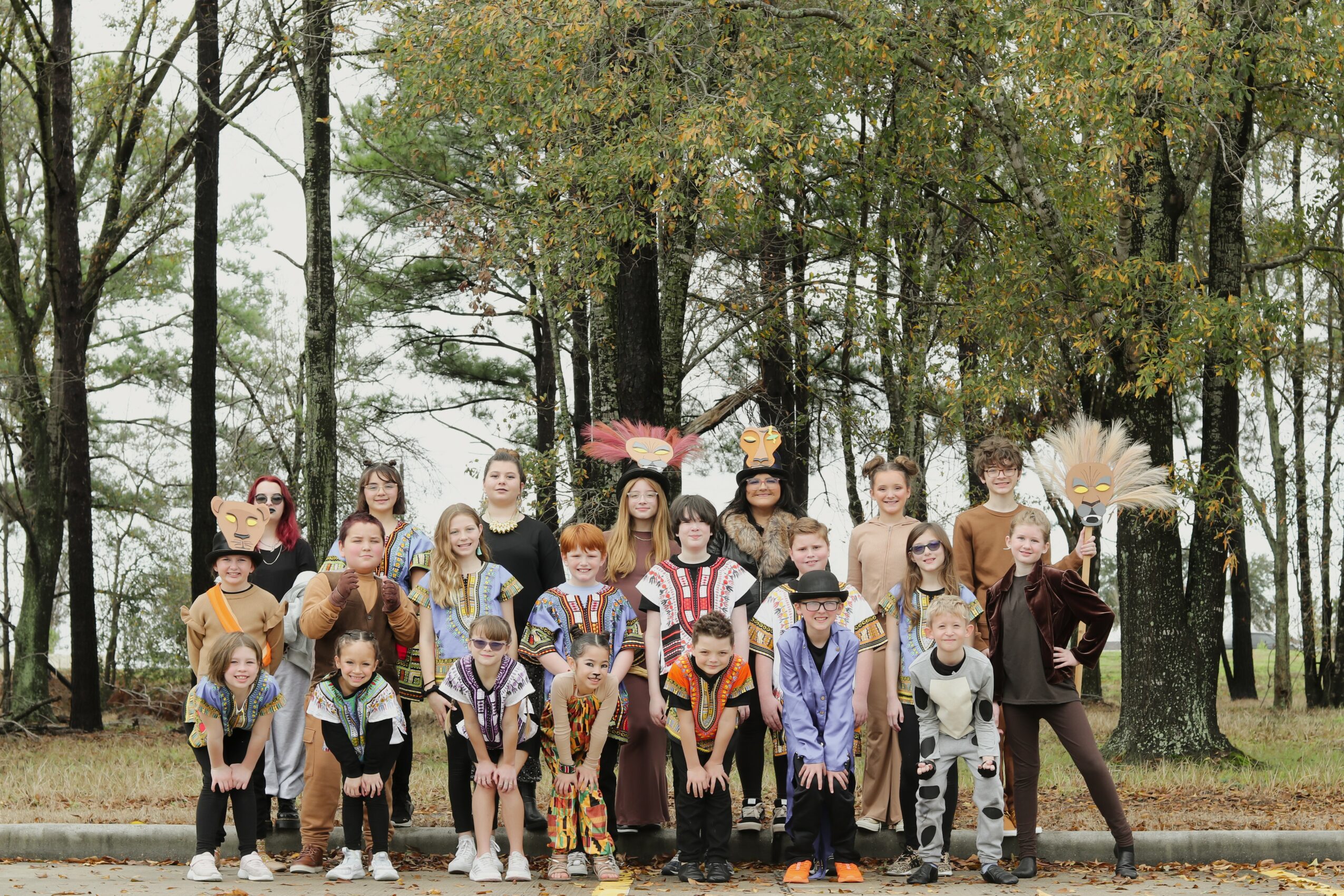 Group of children and adults outdoors in a wooded area, posing for a photo, some wearing costumes or accessories.