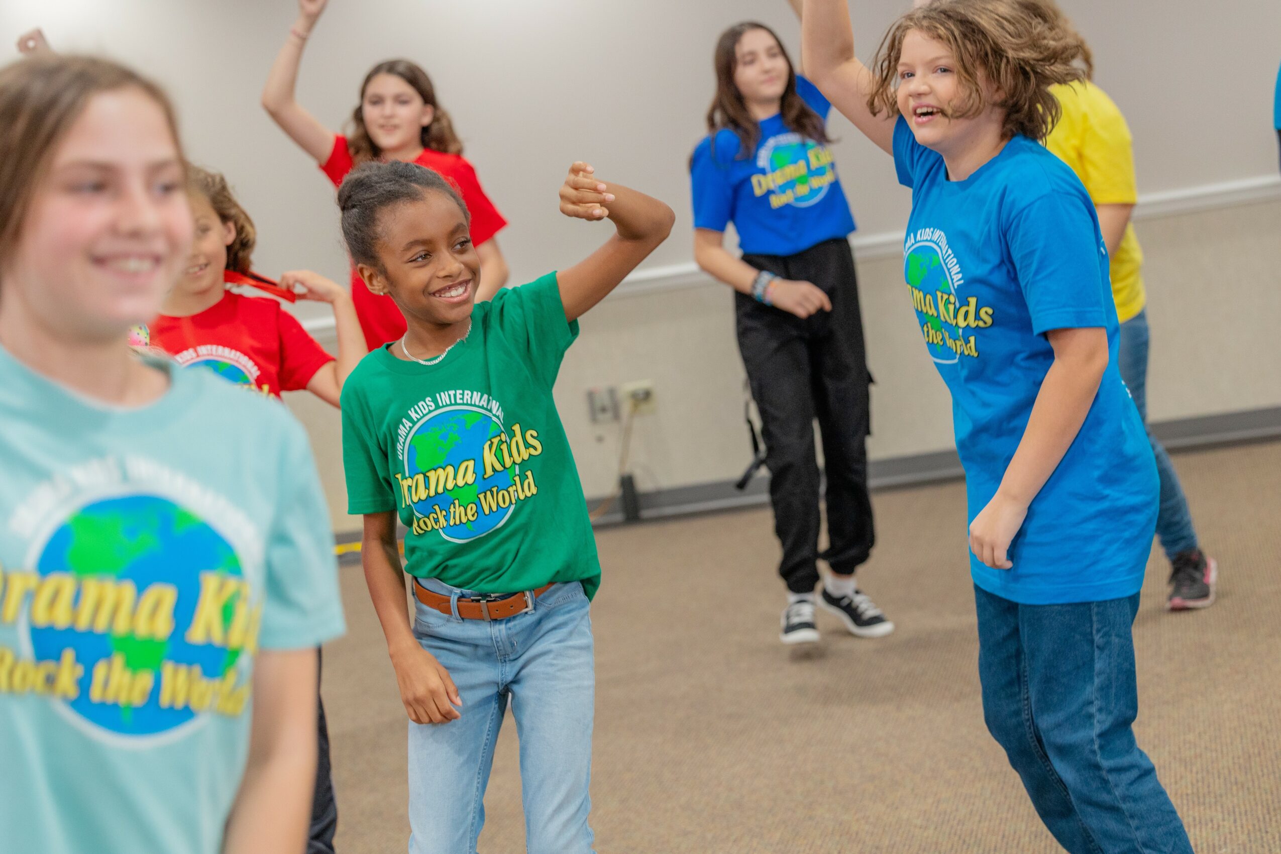 Group of children smiling and raising hands in a classroom or activity room, wearing colorful T-shirts with a logo.