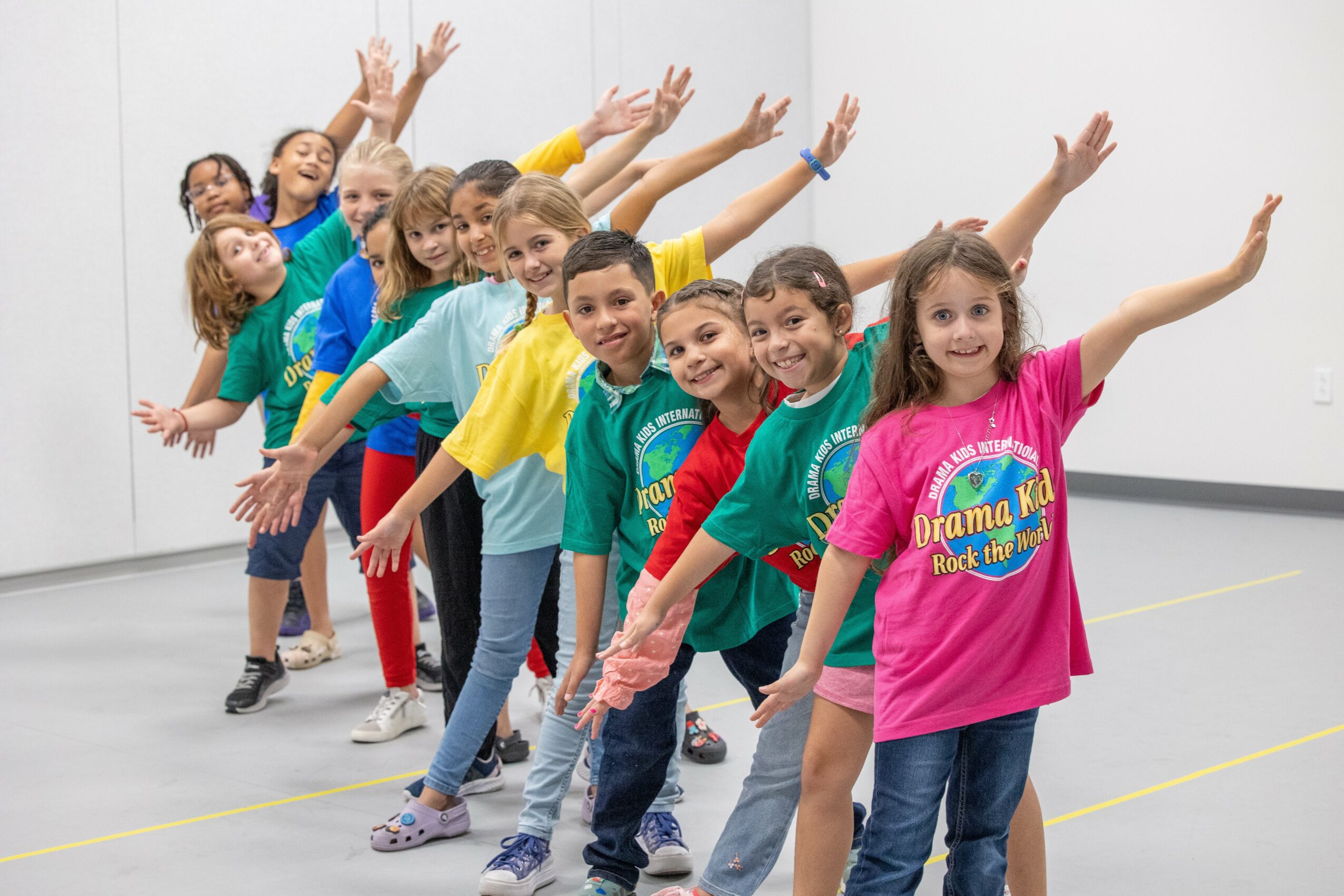 Group of children in colorful shirts standing in a line with arms raised, smiling, in a room with white walls.