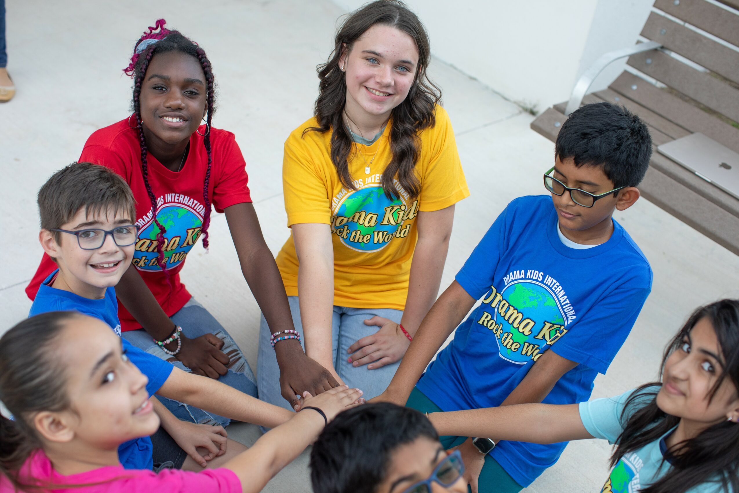 Group of five children smiling and holding hands in a circle indoors, wearing colorful T-shirts.