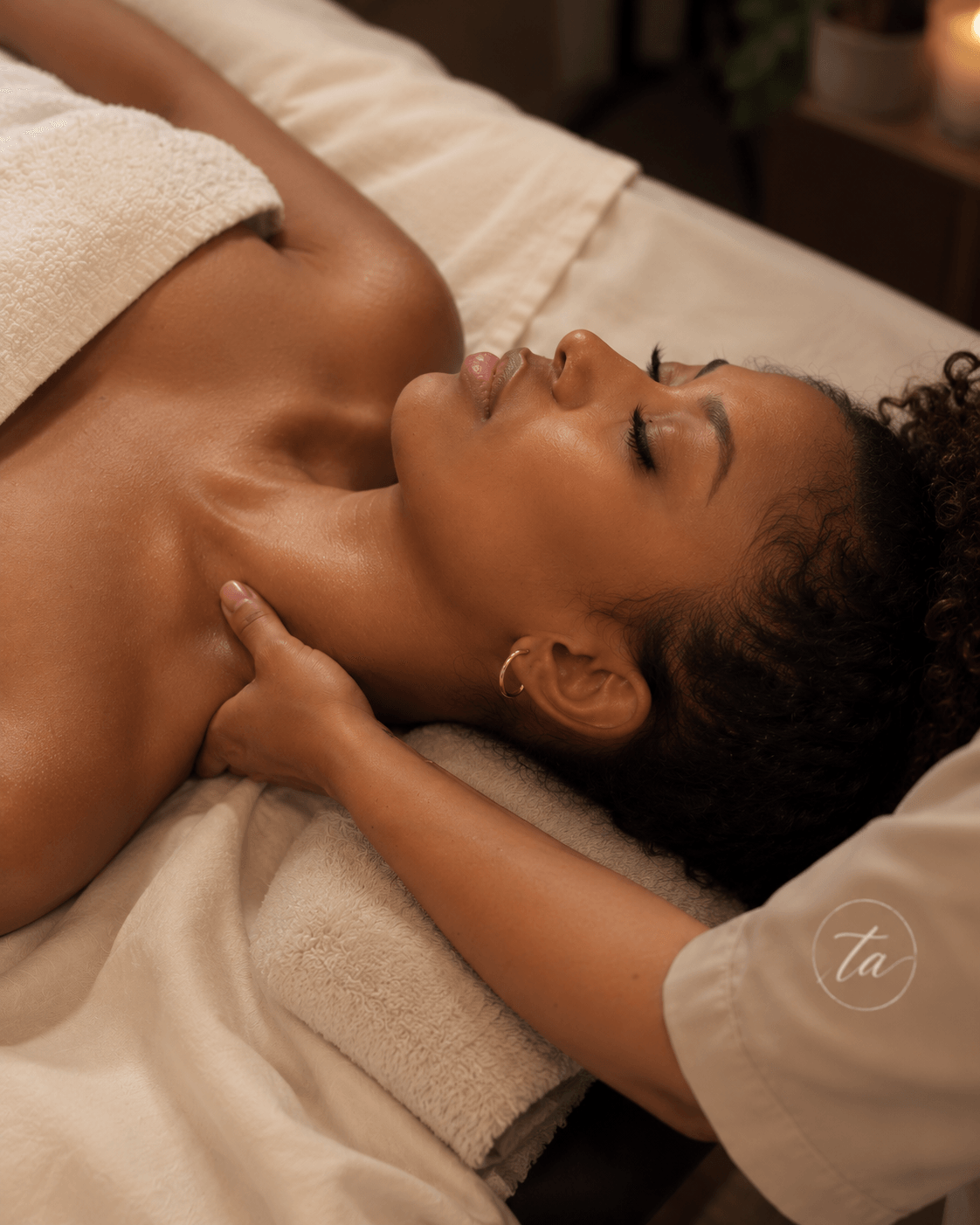 Woman lying on massage table with eyes closed, receiving a massage, wrapped in a towel, in a spa setting.