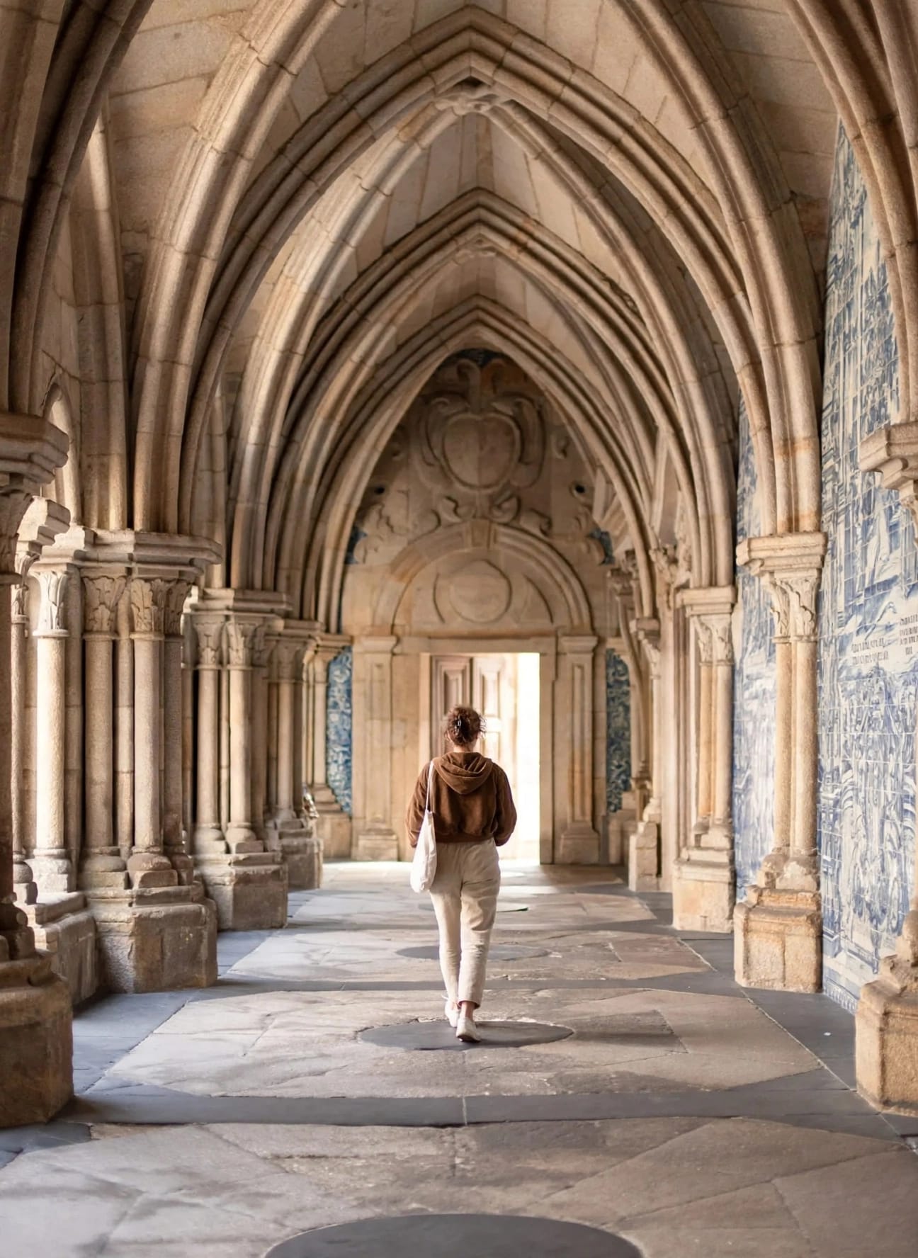 Person walking through a grand, arched stone corridor with intricate architectural details, sunlight streaming in from the end.