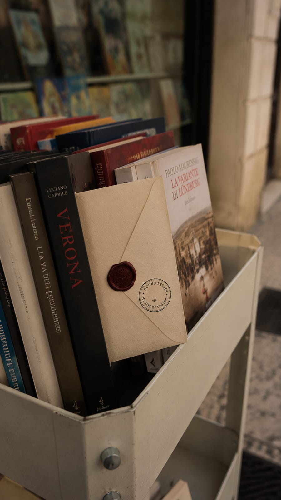 Books in a white bookstand outside a store, with one envelope with a wax seal among them.