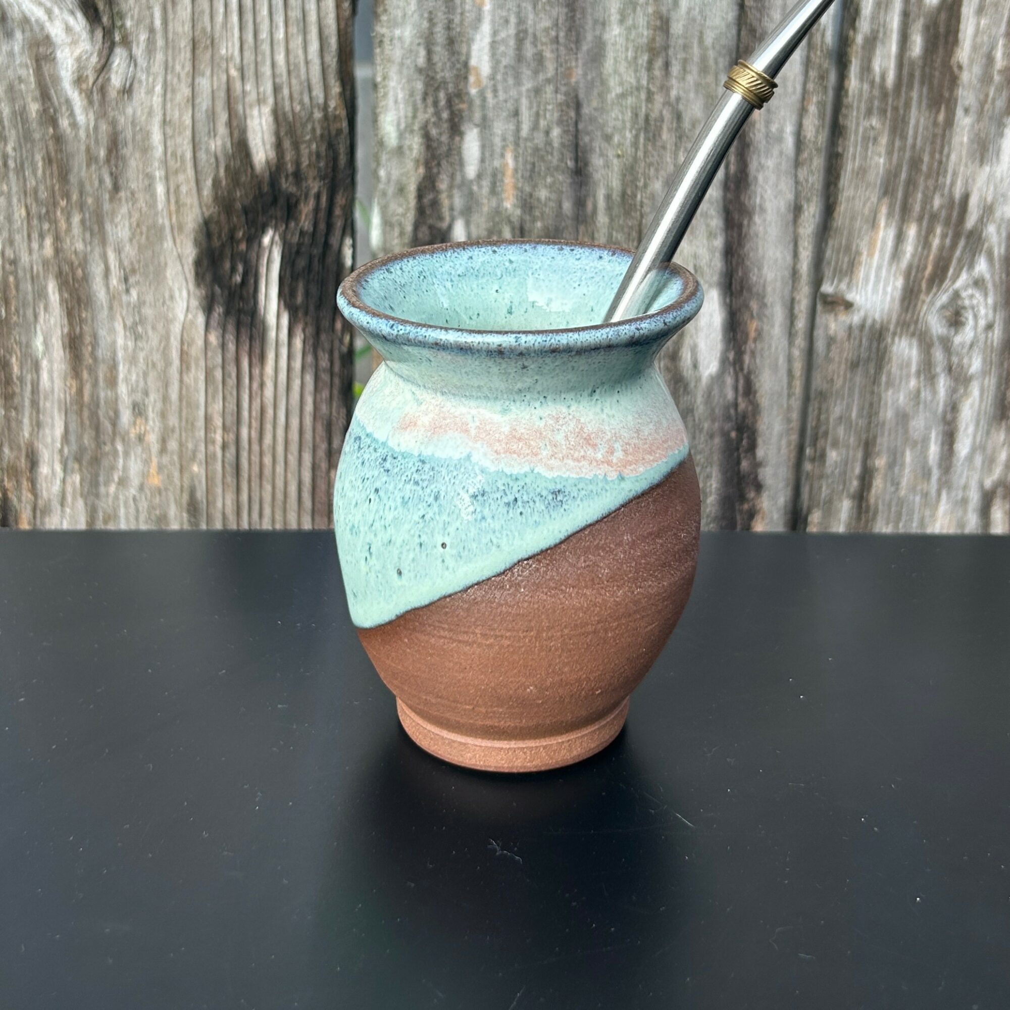 A ceramic cup with a metal straw on a black surface, against a wooden background.