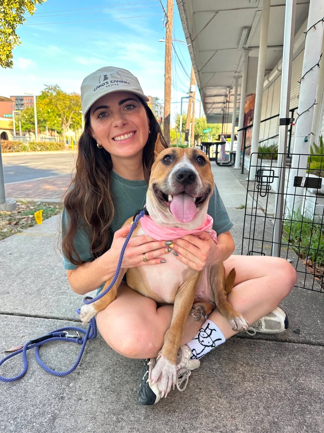 Young woman with long hair and a cap sitting on the sidewalk, holding a happy dog with a pink bandana.