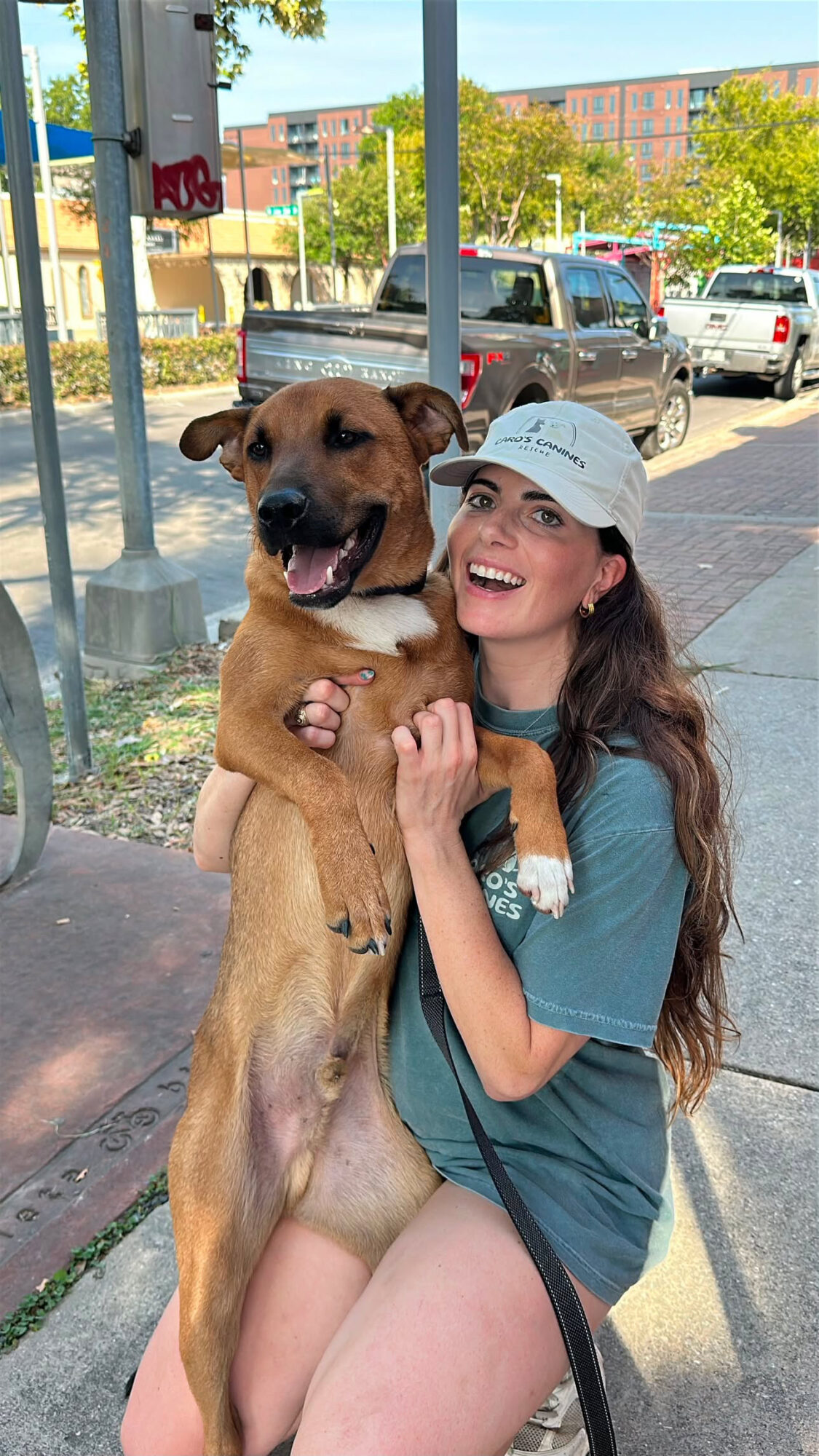 Young woman with long hair, wearing a cap and teal shirt, hugging a large brown dog outdoors on a sidewalk.