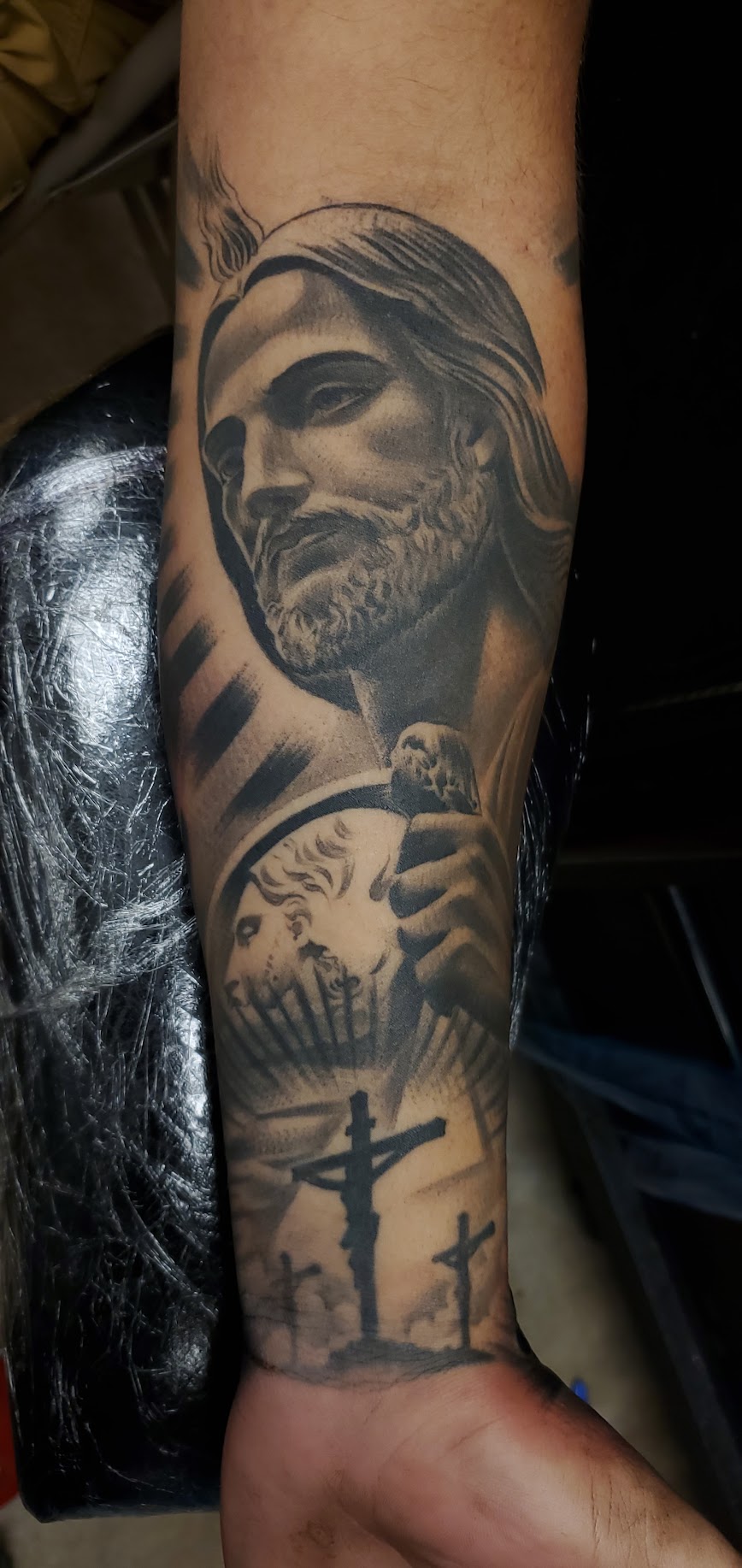Tattoo of a bearded man with long hair holding a cross, with additional crosses and a sunset scene in the background.