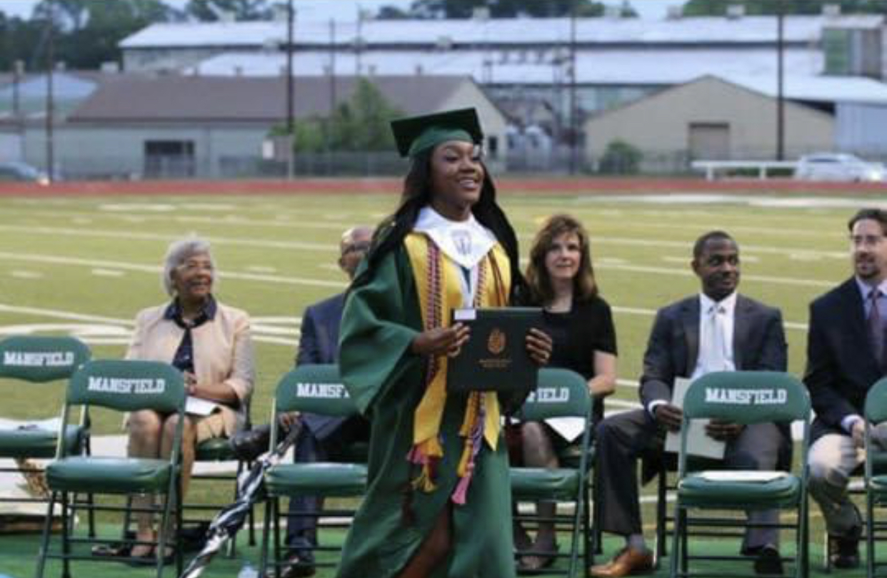 Graduate in cap and gown holding diploma, smiling, with seated people and a track field in background.