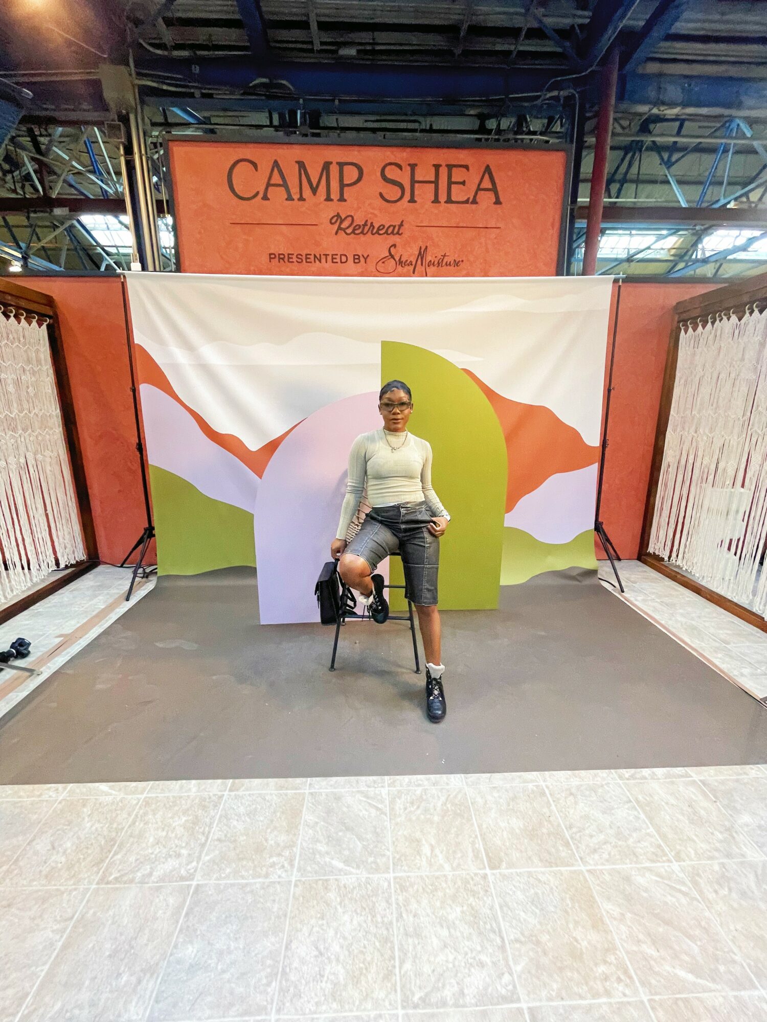 Person sitting on a chair in front of a colorful backdrop with mountains and a large leaf, at Camp Shea event.