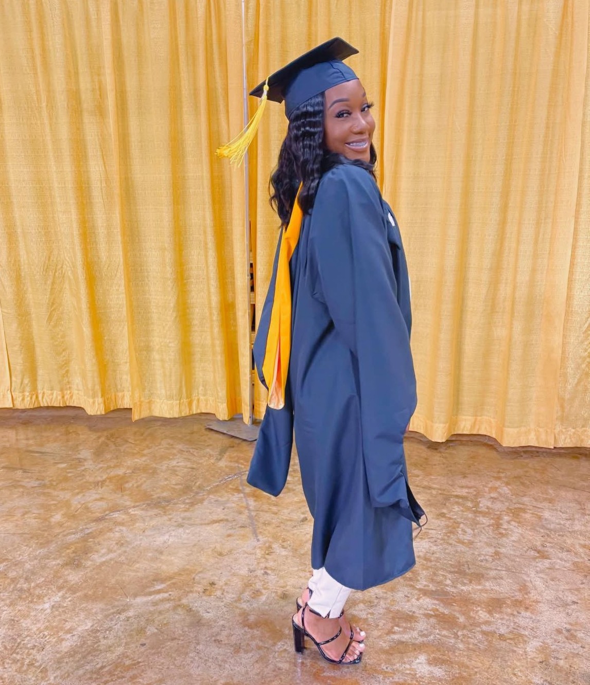 Woman in graduation gown and cap smiling, standing in front of yellow curtain, with heels and white dress visible.
