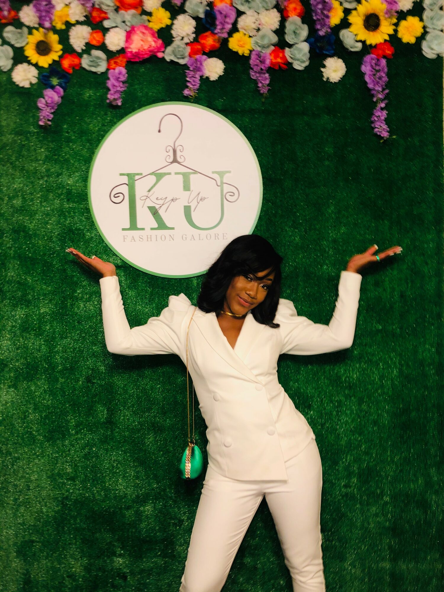 Woman in white suit with arms raised, standing in front of a green wall with flowers and a circular logo.