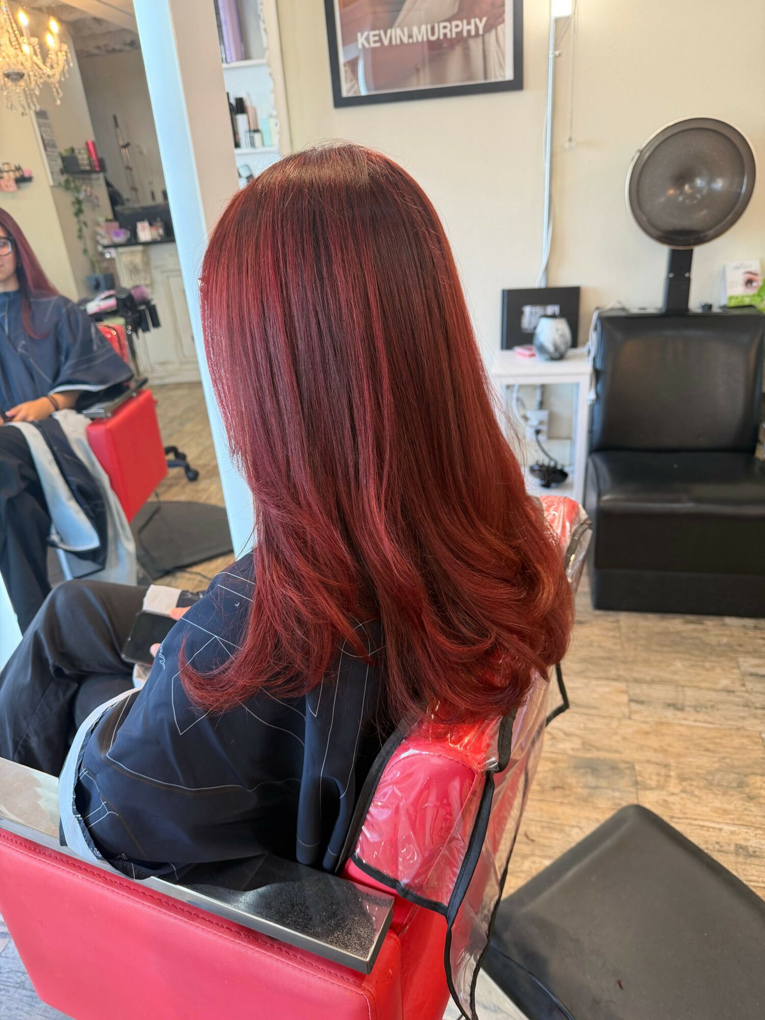 Person with long, red hair sitting in a salon chair, facing away, with salon equipment in the background.