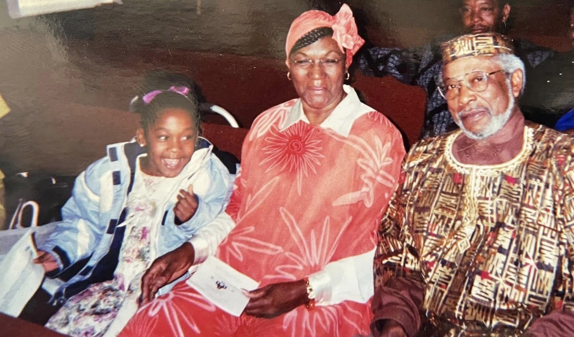 Three people sitting together, smiling, with a dark background. The person in the middle wears a headscarf and red dress. The person on the right wears traditional patterned clothing and glasses. The person on the left is a young girl with a backpack, smiling.