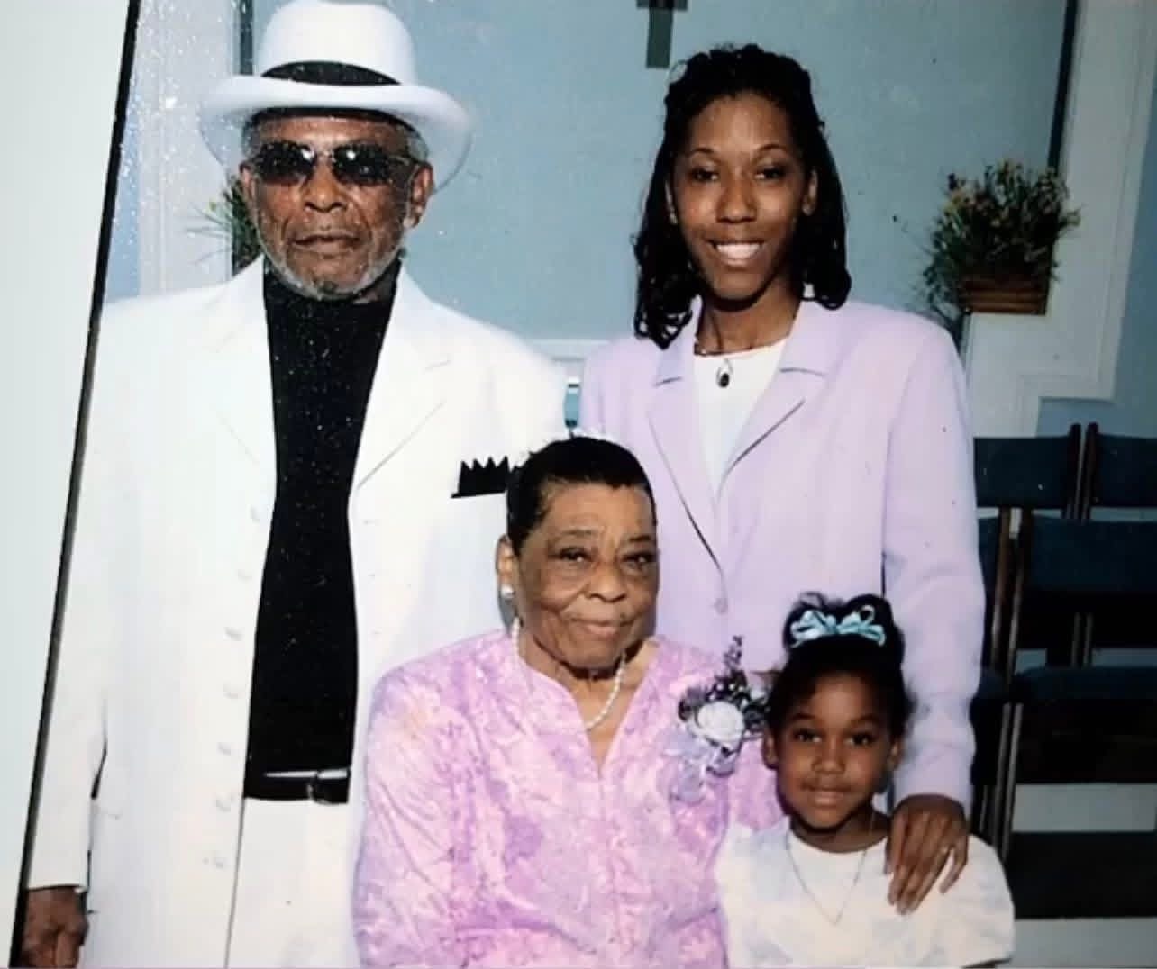 Four people, two adults and two children, pose indoors, smiling, with a staircase and potted plant in the background.