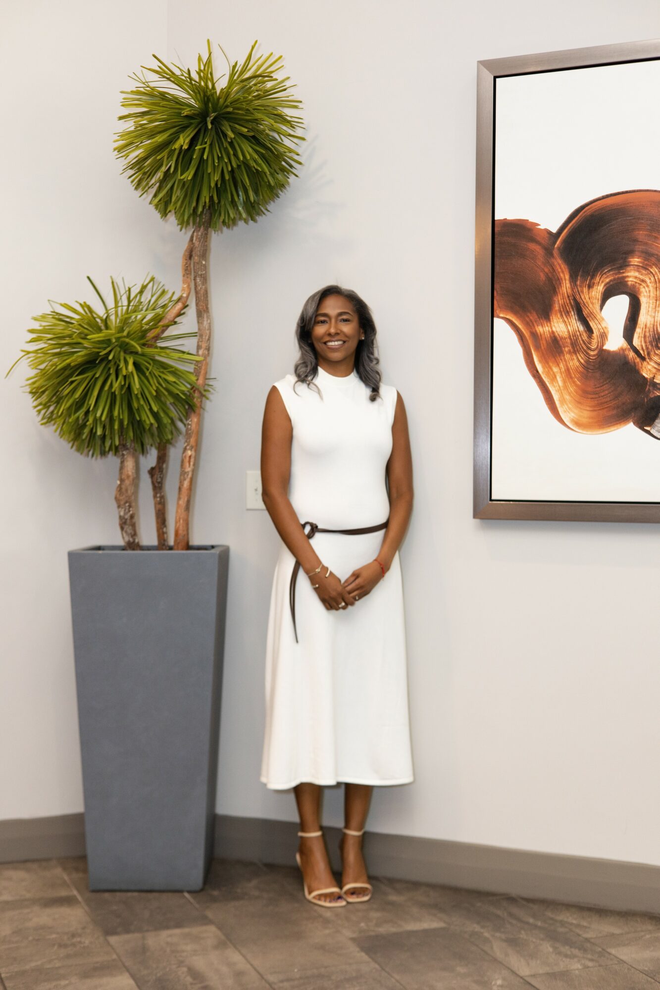 Woman in white dress standing next to two tall potted plants and abstract artwork on wall.