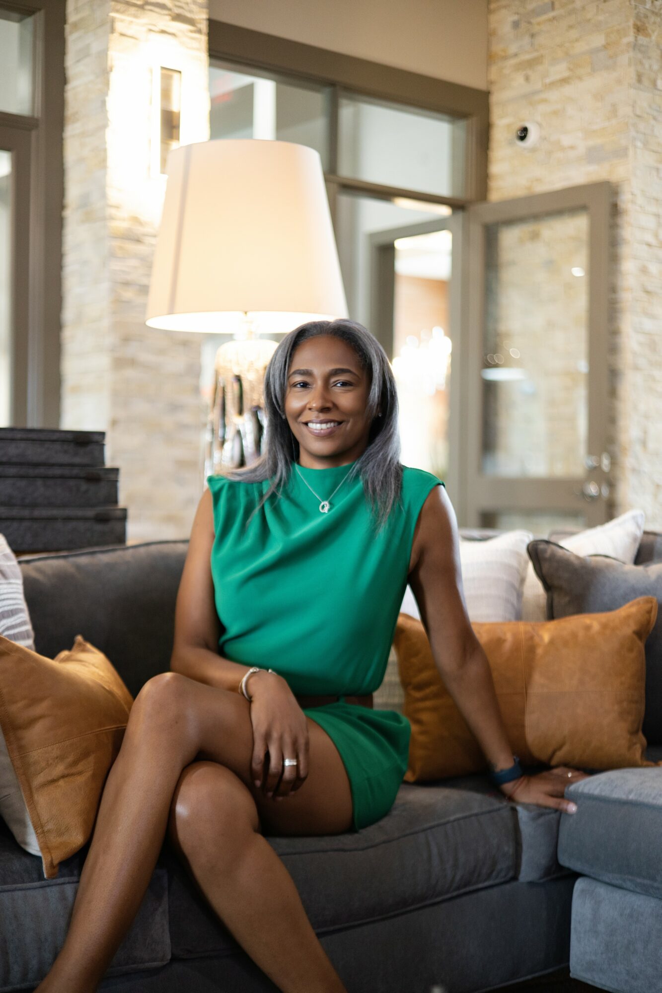 Smiling woman in green dress sitting on a sofa in a modern living room with large windows and a lamp.