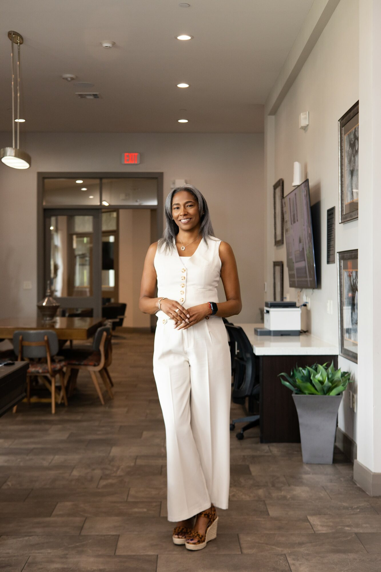 Woman in white sleeveless outfit standing in a modern indoor space with framed pictures and a potted plant, smiling.
