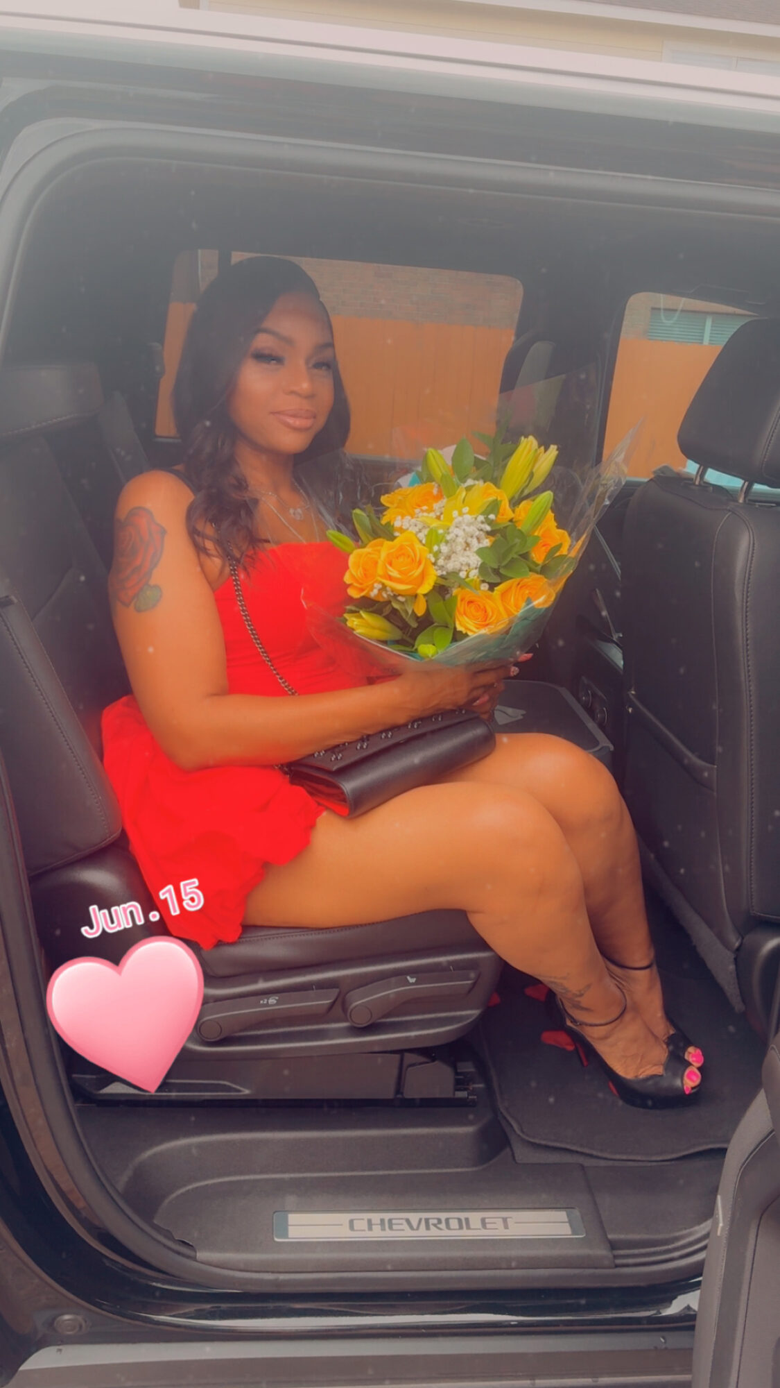 Woman in red dress holding a bouquet of yellow flowers, sitting in a car seat, with a pink heart sticker and date overlay.