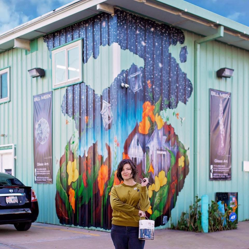 Woman standing in front of a colorful mural on a building, with a mountain and sky scene, holding a bag.