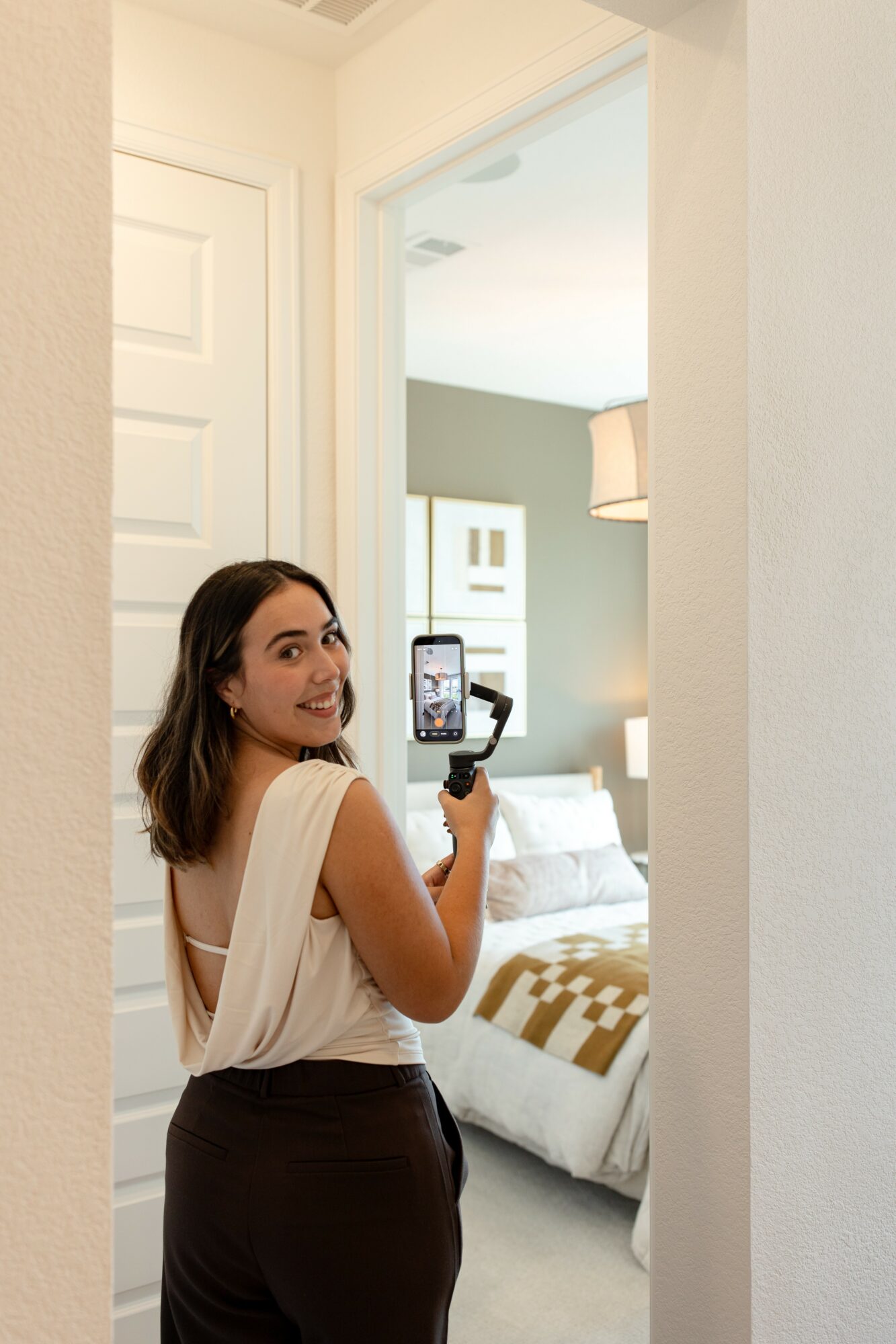 Young woman with shoulder-length hair smiling and taking a selfie in a bedroom doorway.