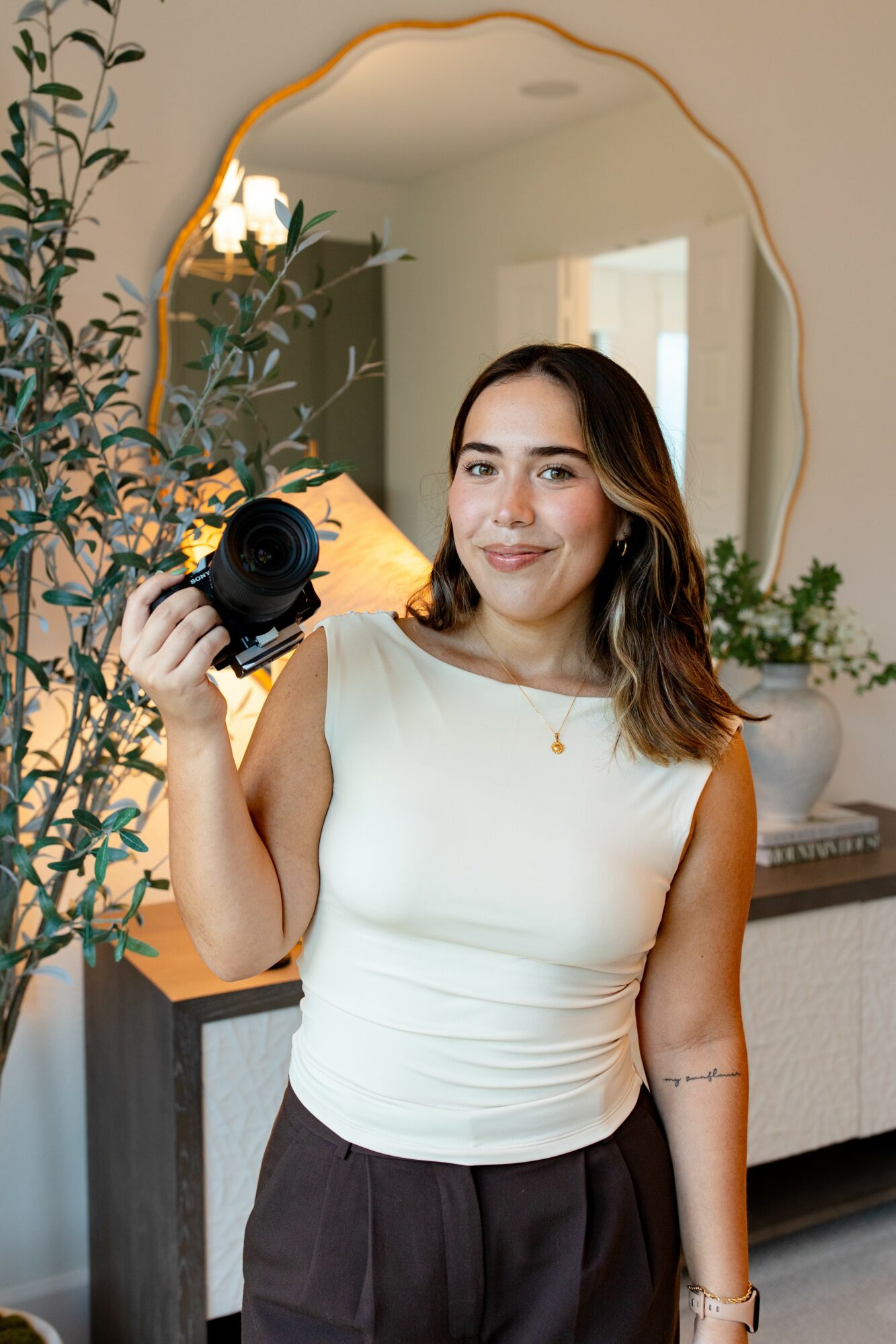 Woman with shoulder-length hair holding a camera, smiling indoors near a mirror and plants.