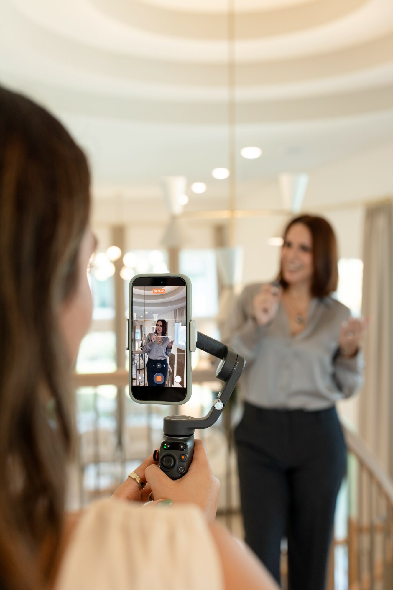 Person recording a woman speaking in a bright indoor space with a smartphone mounted on a stabilizer.