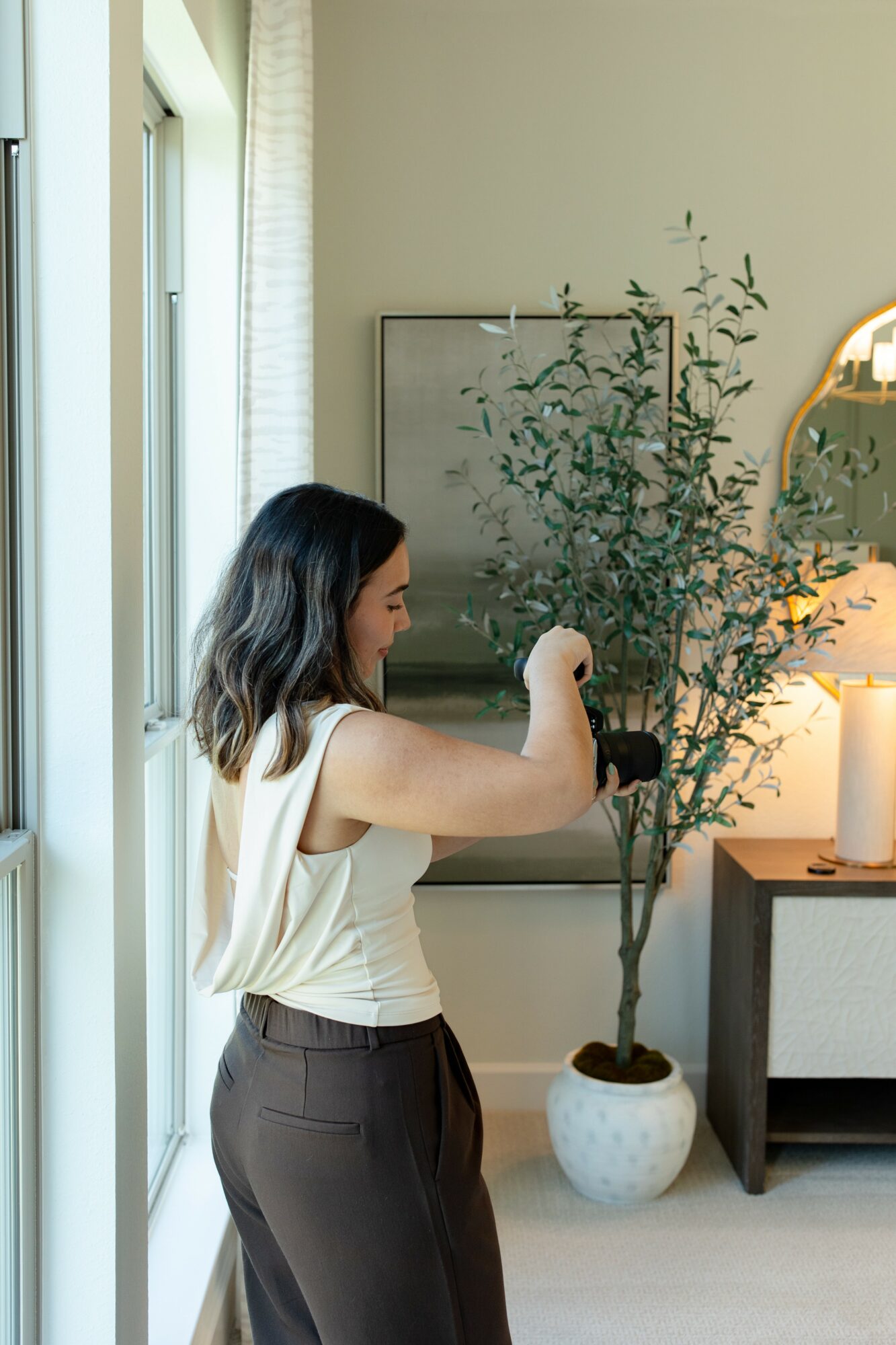 Woman with shoulder-length hair arranging a potted plant indoors near a window.