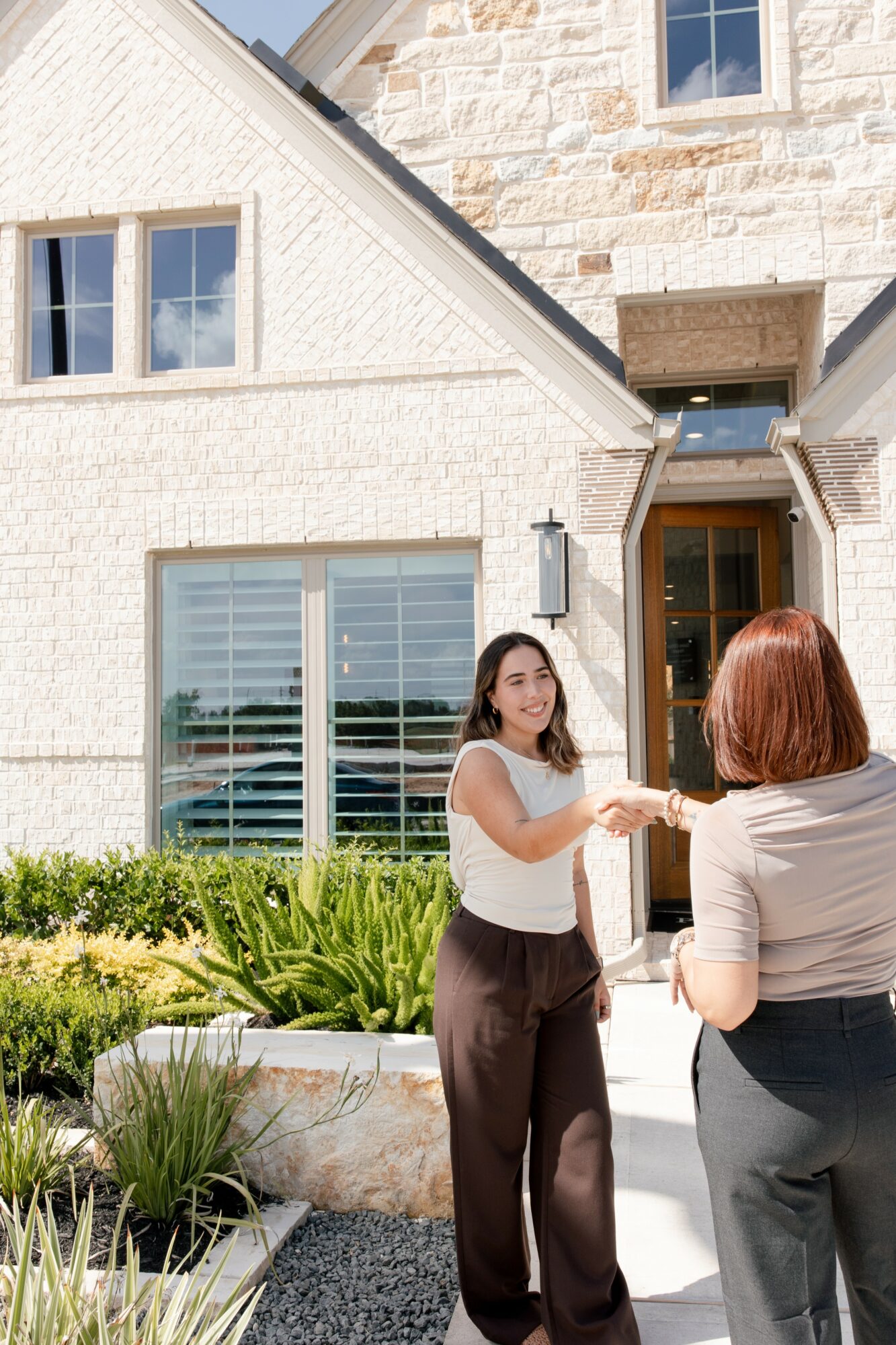 Two women shaking hands outside a white house with large windows and a garden, sunny day.
