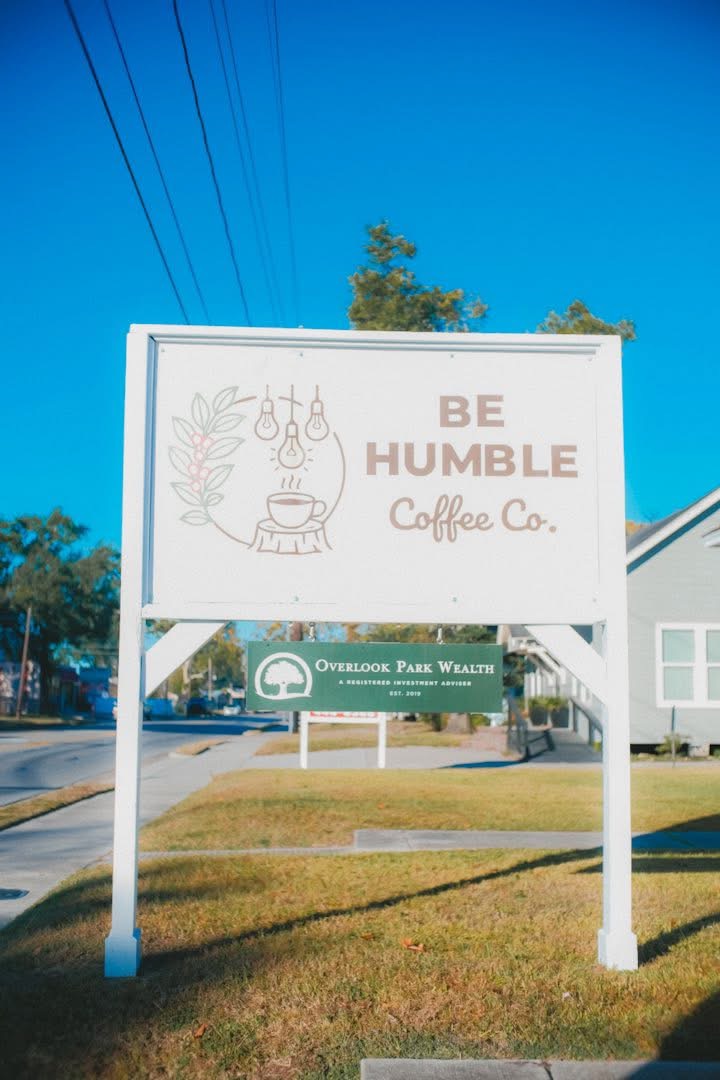 Sign with text 'BE HUMBLE Coffee Co.' and a decorative illustration, against a blue sky with trees and power lines.