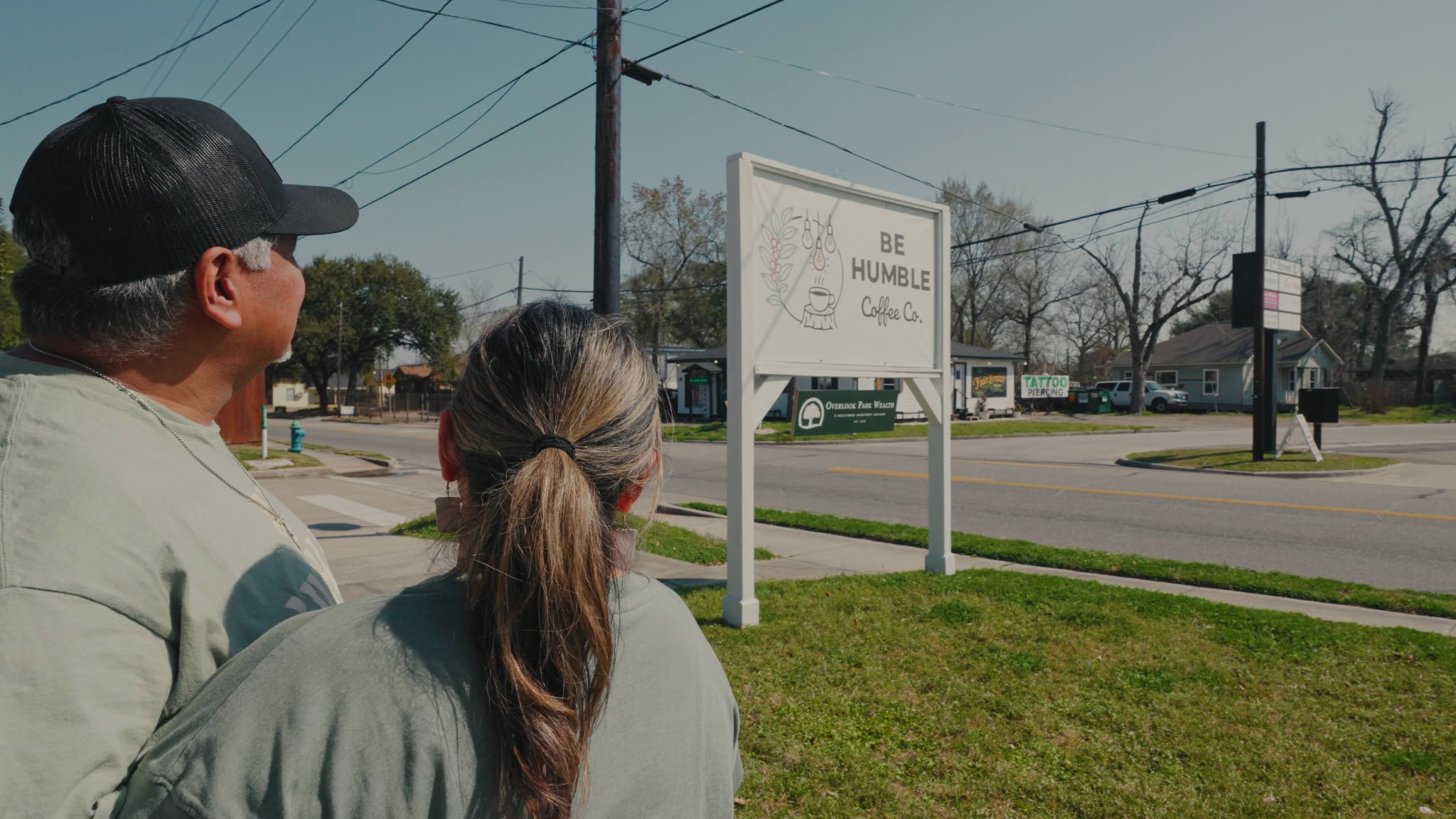 Two people stand outdoors near a sign and a street, with trees and power lines in the background.
