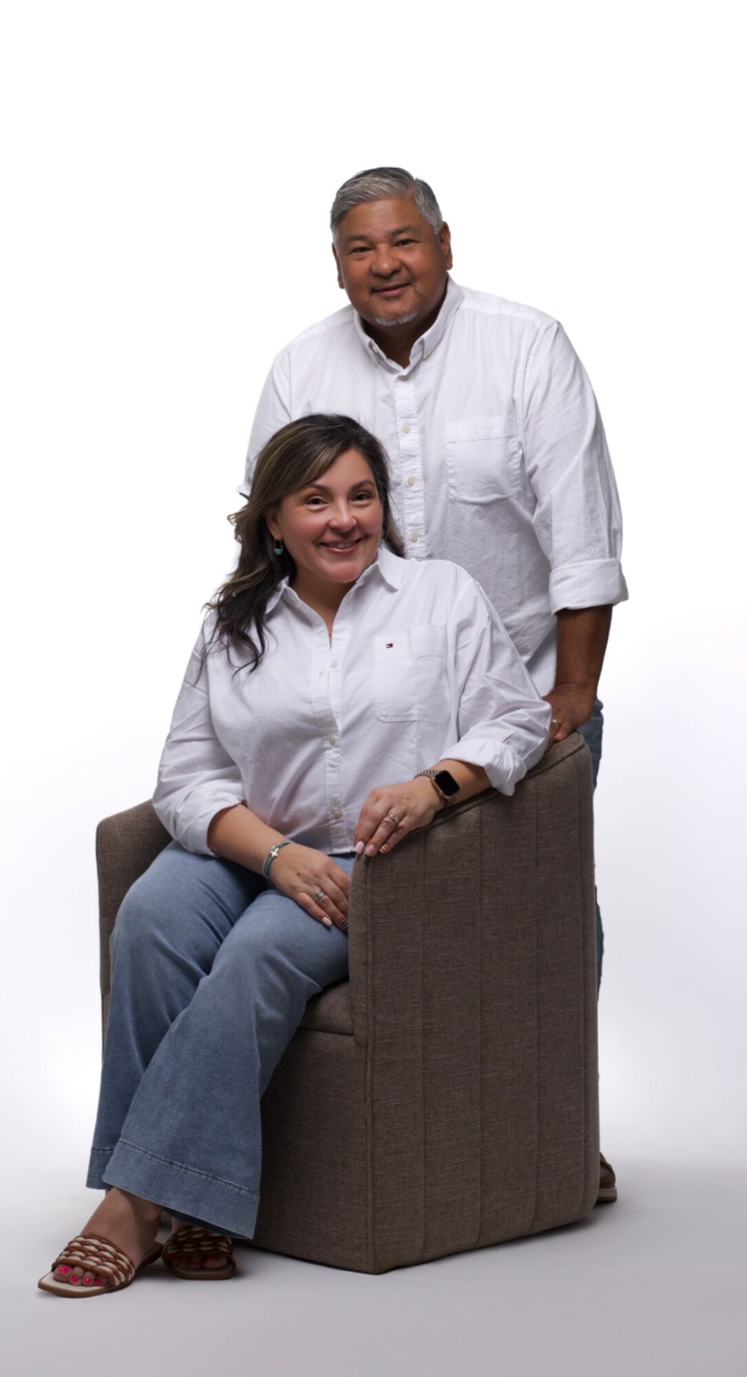 A man and woman pose together, the woman sitting on a chair and the man standing behind her, both smiling.