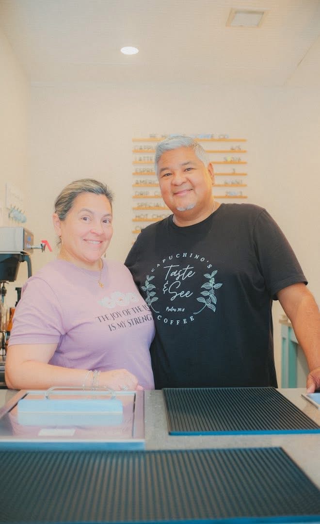 Two people smiling indoors, one wearing a pink shirt and the other a black shirt, standing behind a counter.