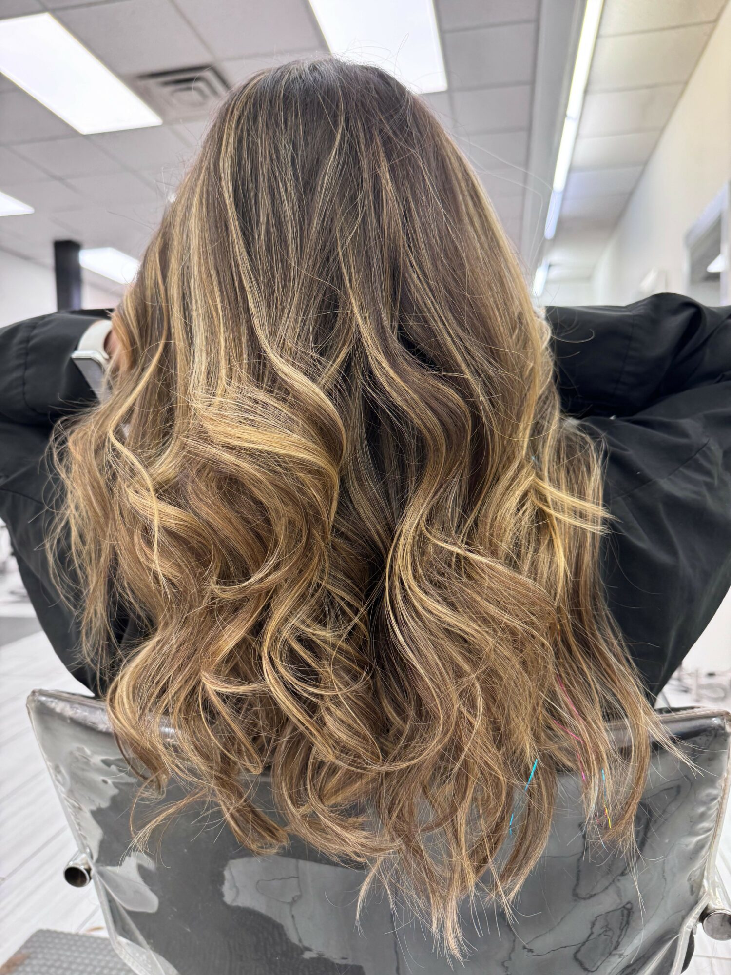 Person with long, wavy, blonde hair sitting in a salon chair, facing away, with a black cape around shoulders.