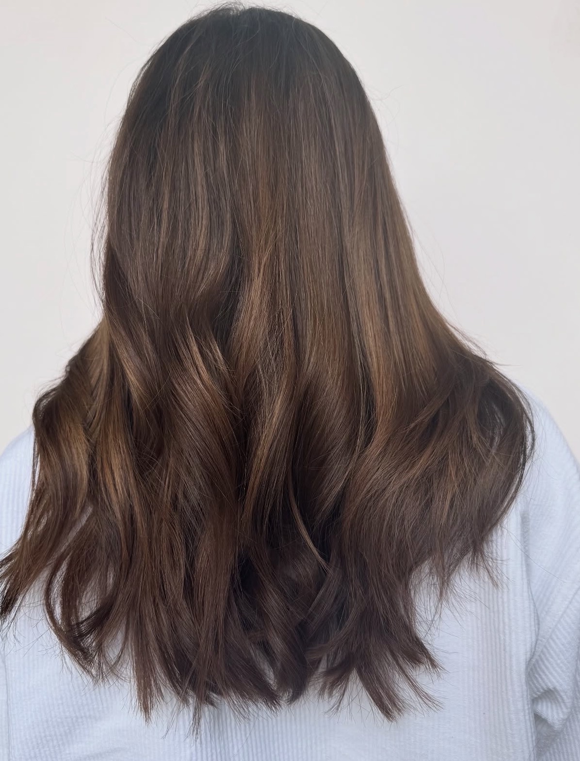 Back view of a person with long, wavy brown hair, wearing a light-colored top.