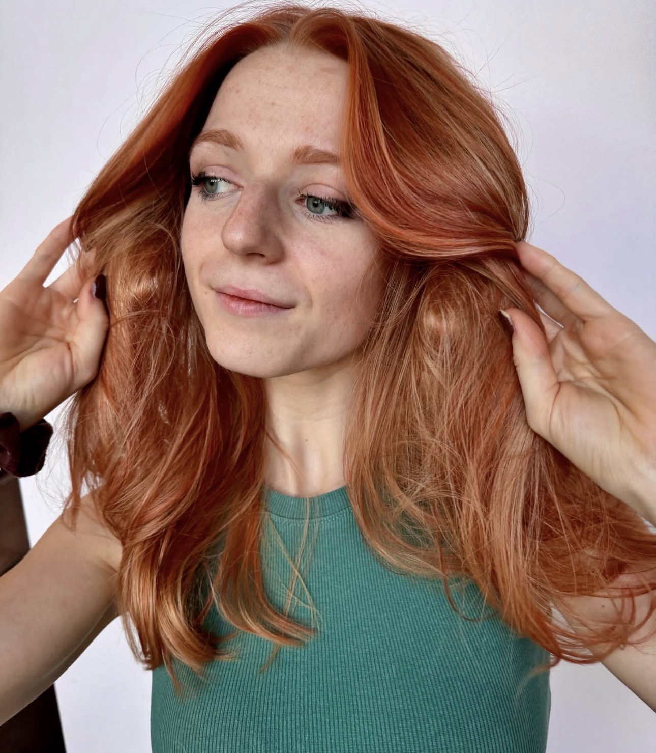 Woman with long, wavy red hair looking to the side, touching her hair, wearing a teal top, neutral background.
