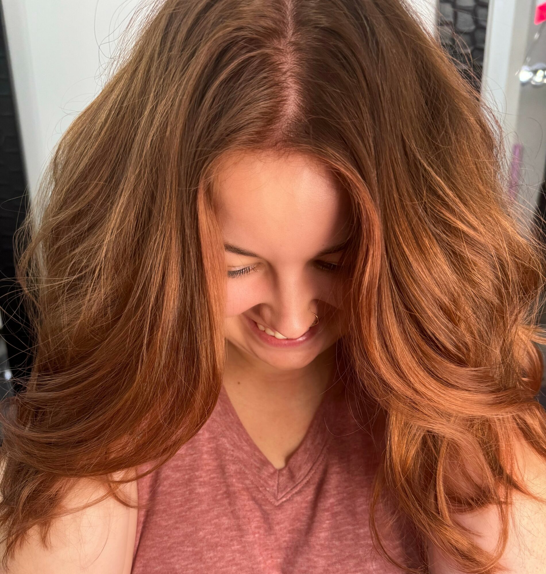 Woman with long, wavy red hair looking down and smiling, wearing a pink top, indoors.