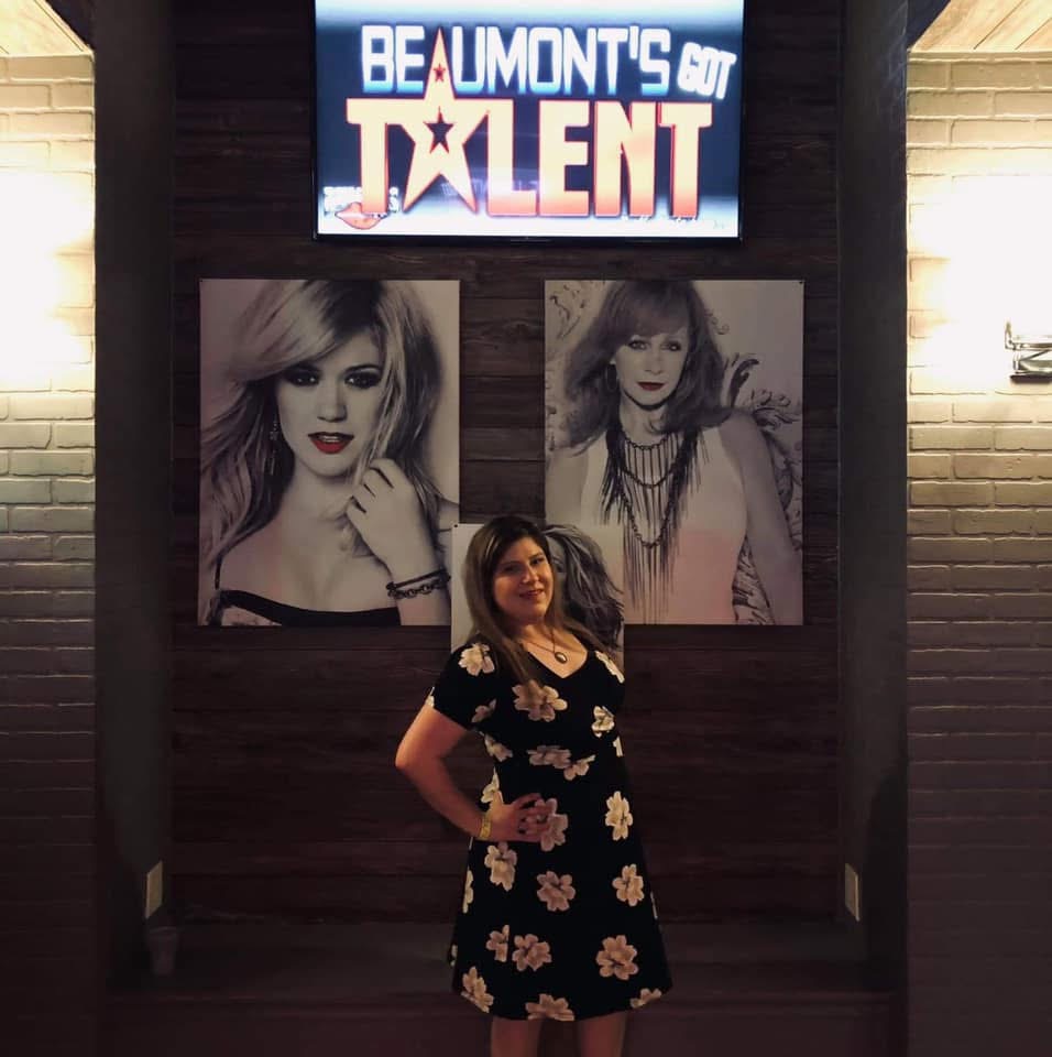 Woman in a black dress with floral pattern standing in front of two large black-and-white portraits, with a sign above reading 'BEAUMONT'S Got TALENT'.